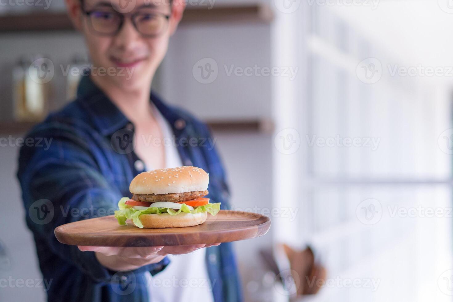 Young man wearing glasses smiling, looking at the camera, and holding a homemade hamburger on a wooden dish with blur background. Space for text. Selective focus. Concept of cooking photo