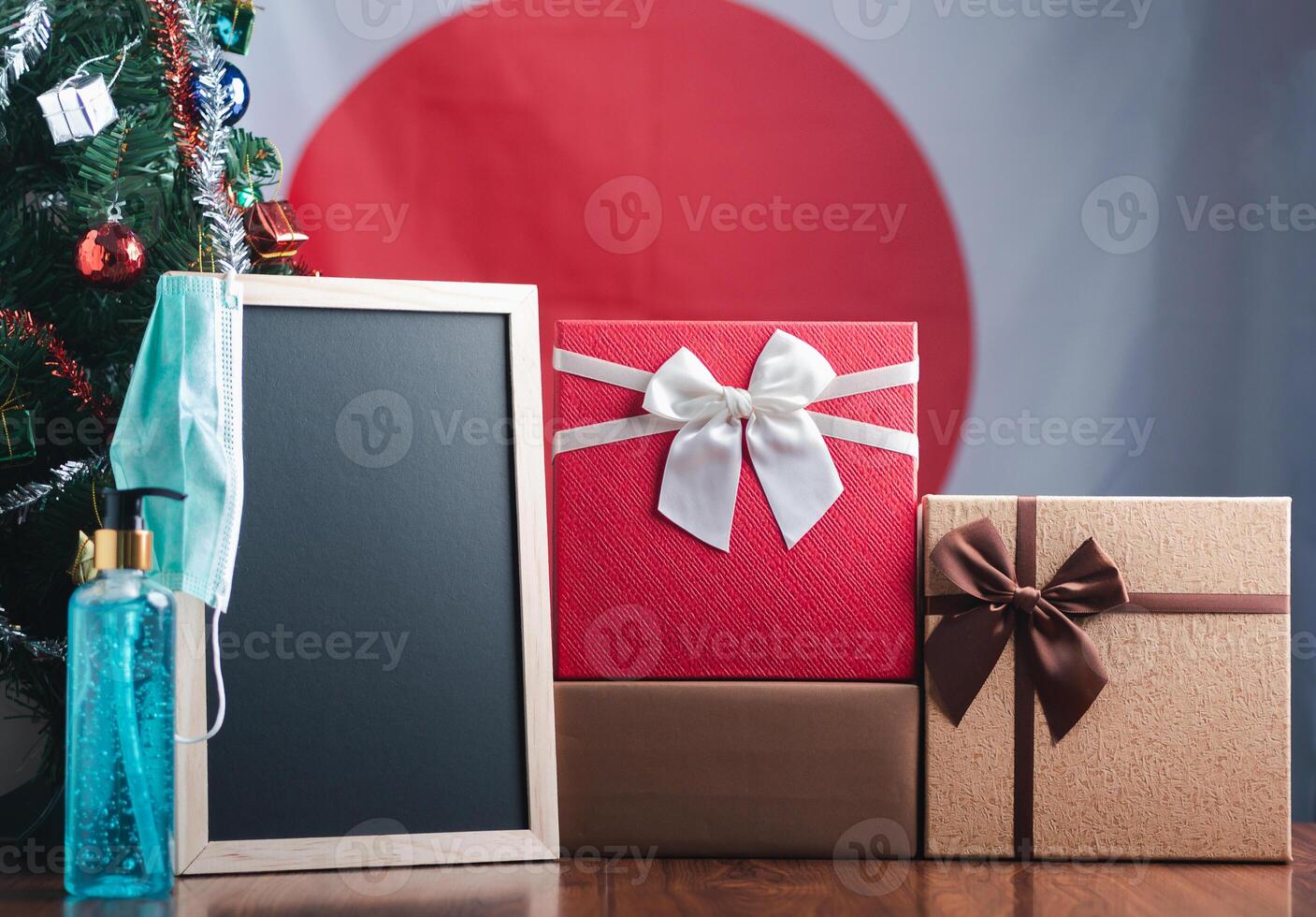 Small blackboard, mask, alcohol gel, and gift boxes on wooden table with a Christmas tree and Japan flag in the background. Space for text. Concept of Christmas and new year festival photo