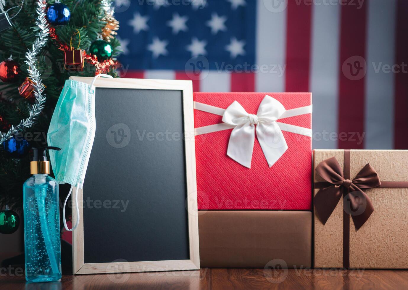 Small blackboard, mask, alcohol gel, and gift boxes on wooden table with a Christmas tree and American flag in the background. Space for text. Concept of Christmas and new year festival photo