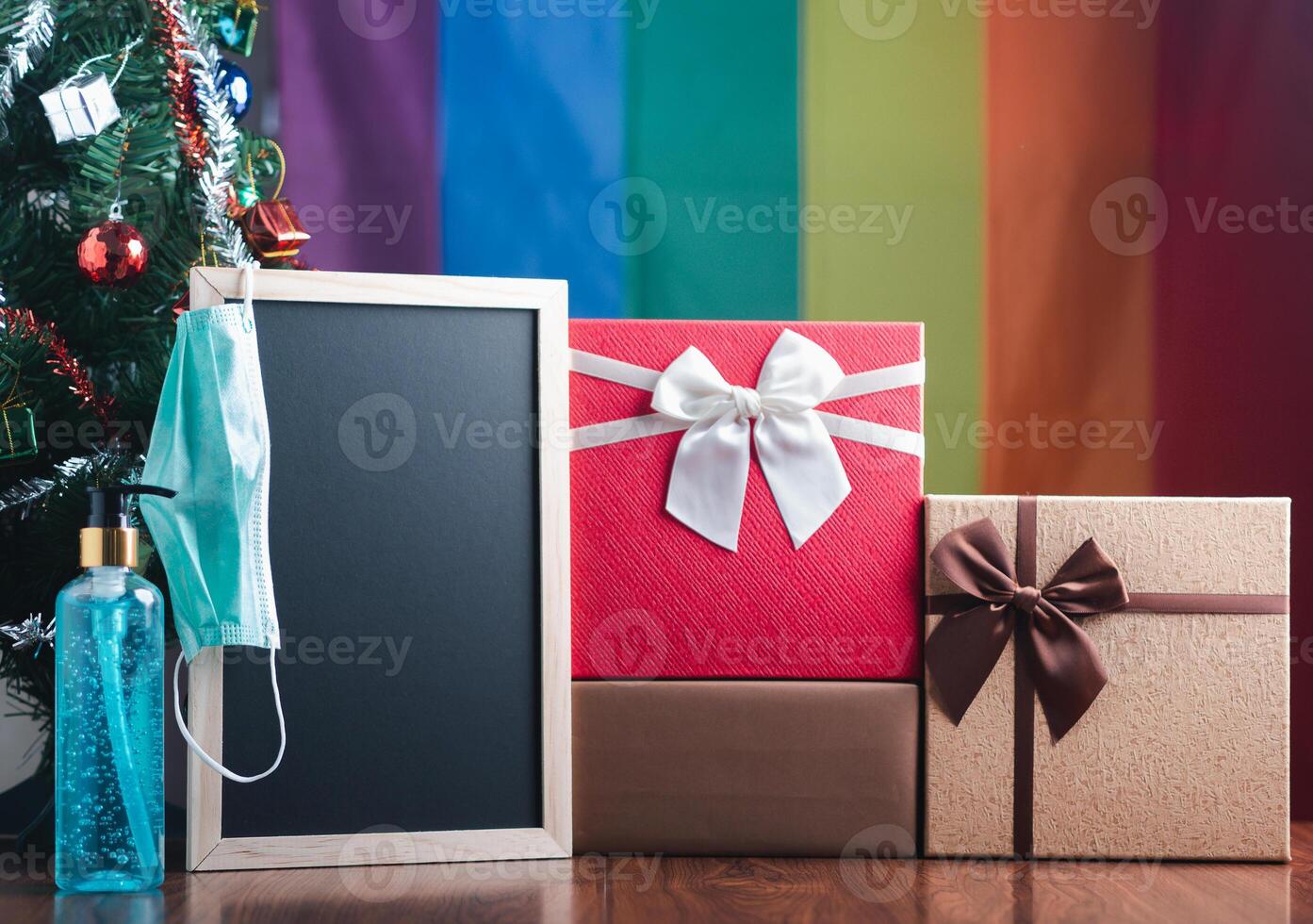 Small blackboard, mask, alcohol gel, and gift boxes on wooden table with a Christmas tree and LGBT flag in the background. Space for text. Concept of Christmas and new year festival photo