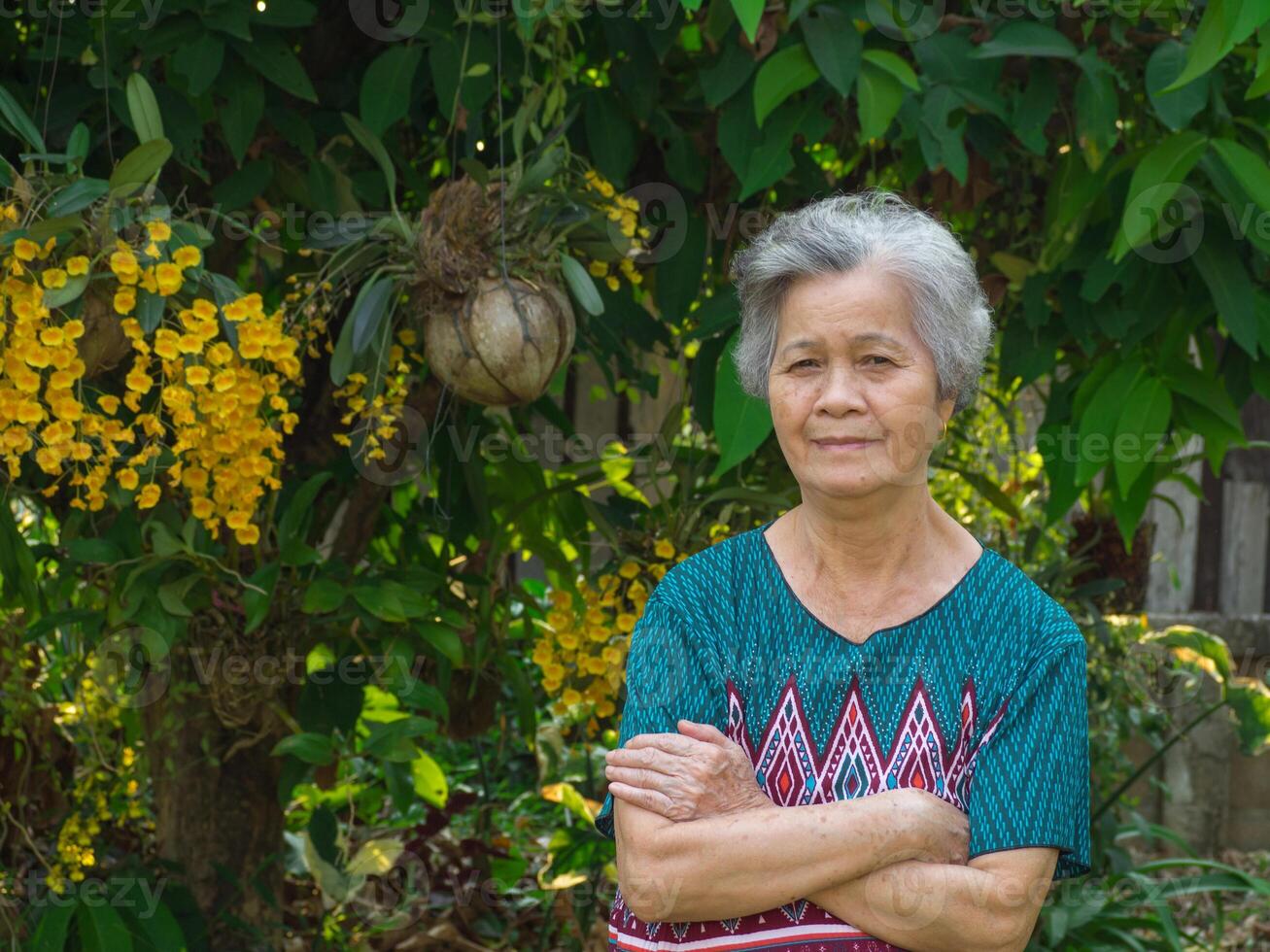 Senior woman smiling, arms crossed and looking at the camera while standing beside beautiful yellow orchids in a garden. photo