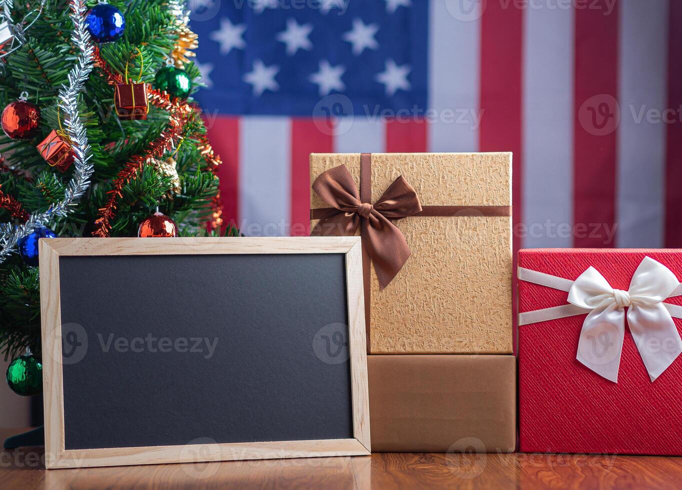 Small blackboard and gift boxes on wooden table with a Christmas tree and American flag in the background. Space for text. Concept of Christmas and new year festival photo