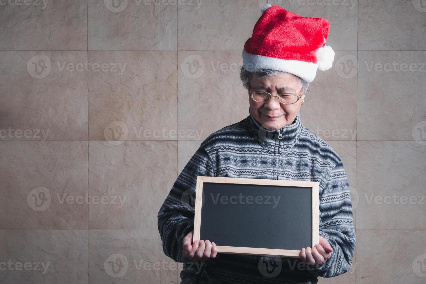 Portrait of an elderly Asian woman wearing a Santa Claus hat, holding a small blackboard and looking down while standing with a light yellow background. Concept of aged people and festival photo