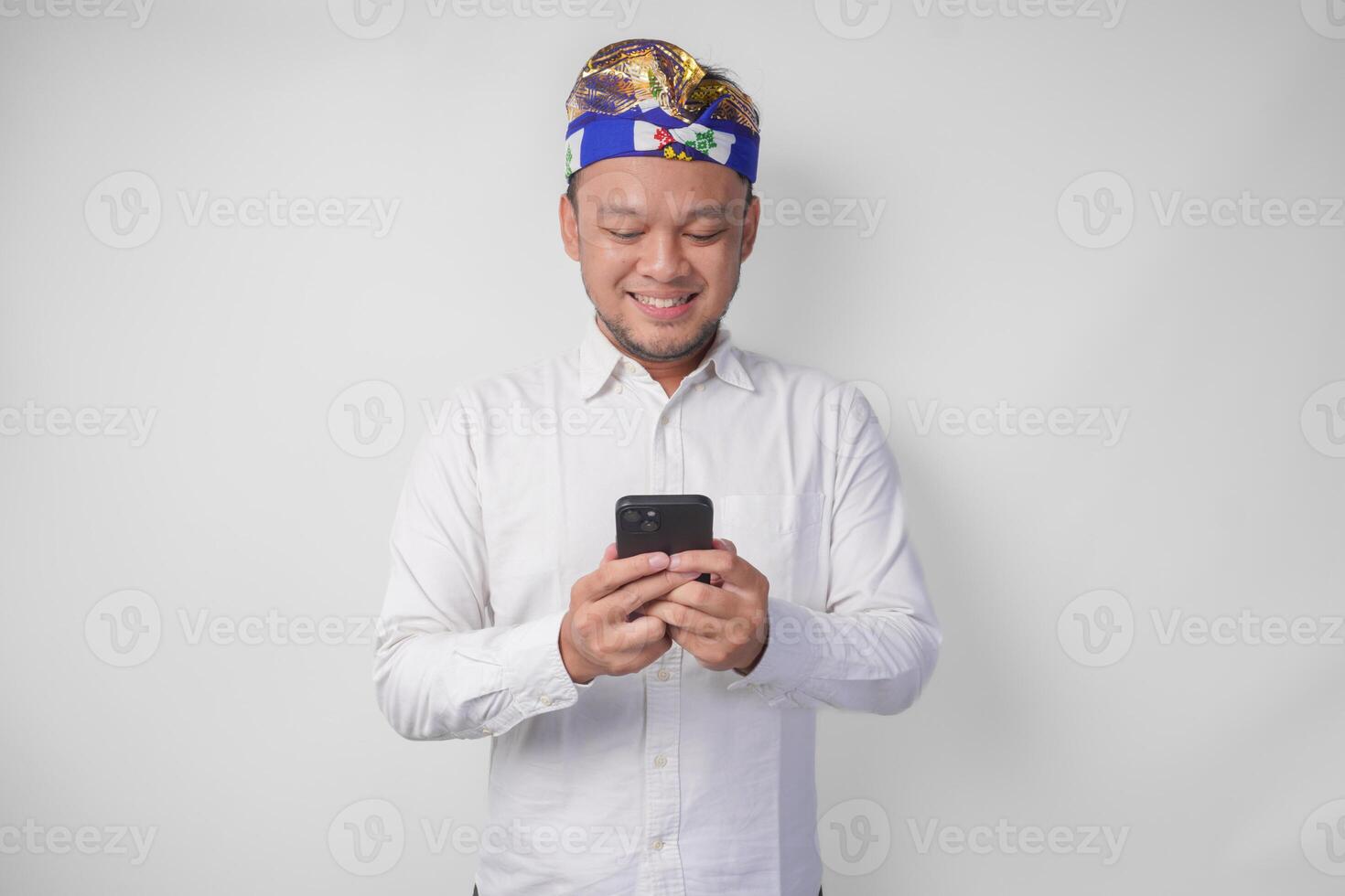Excited Balinese man wearing white shirt and traditional headdress smiling happily while using smartphone, replying message, reading news photo