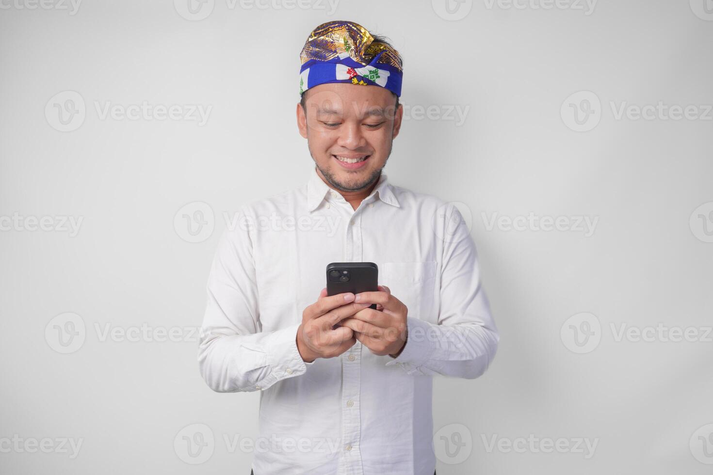 emocionado balinés hombre vistiendo blanco camisa y tradicional tocado sonriente felizmente mientras utilizando teléfono inteligente, respondiendo mensaje, leyendo Noticias foto