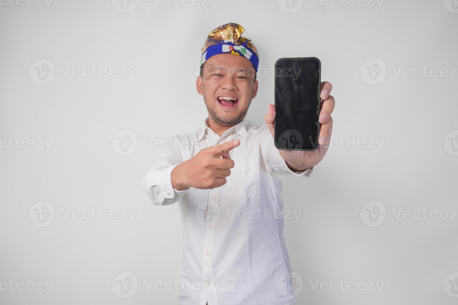 Smiling young Balinese man wearing white shirt and traditional headdress pointing to his smartphone, presenting blank screen copy space photo