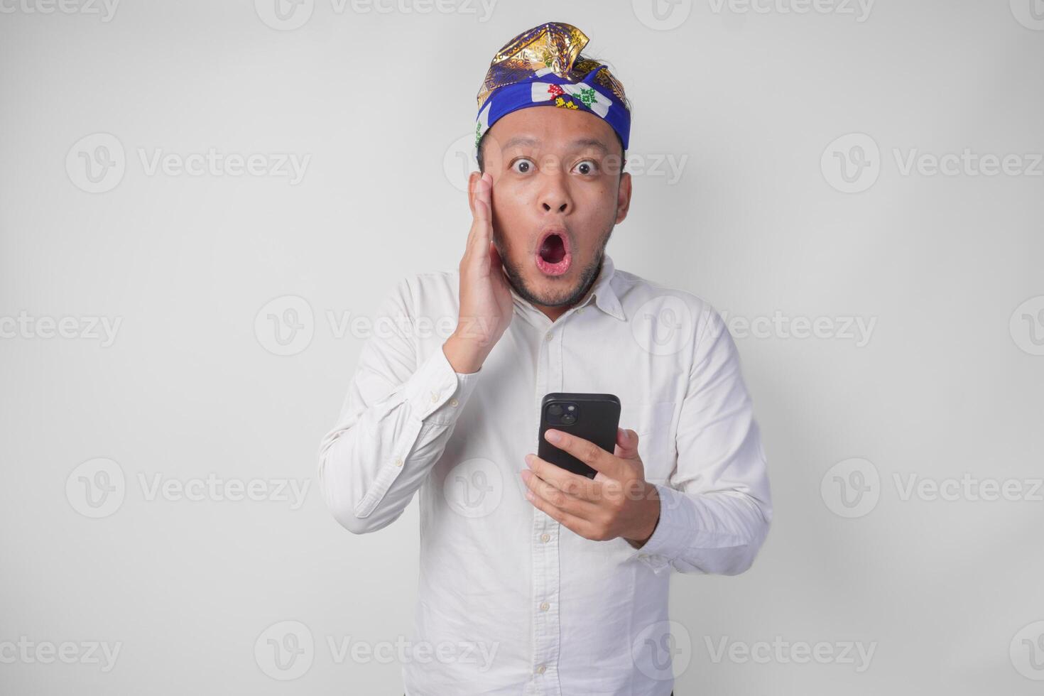 Young Balinese man wearing white shirt and traditional headdress showing shocked expression on face while holding smartphone, surprised after reading news or gossip, isolated by white background photo