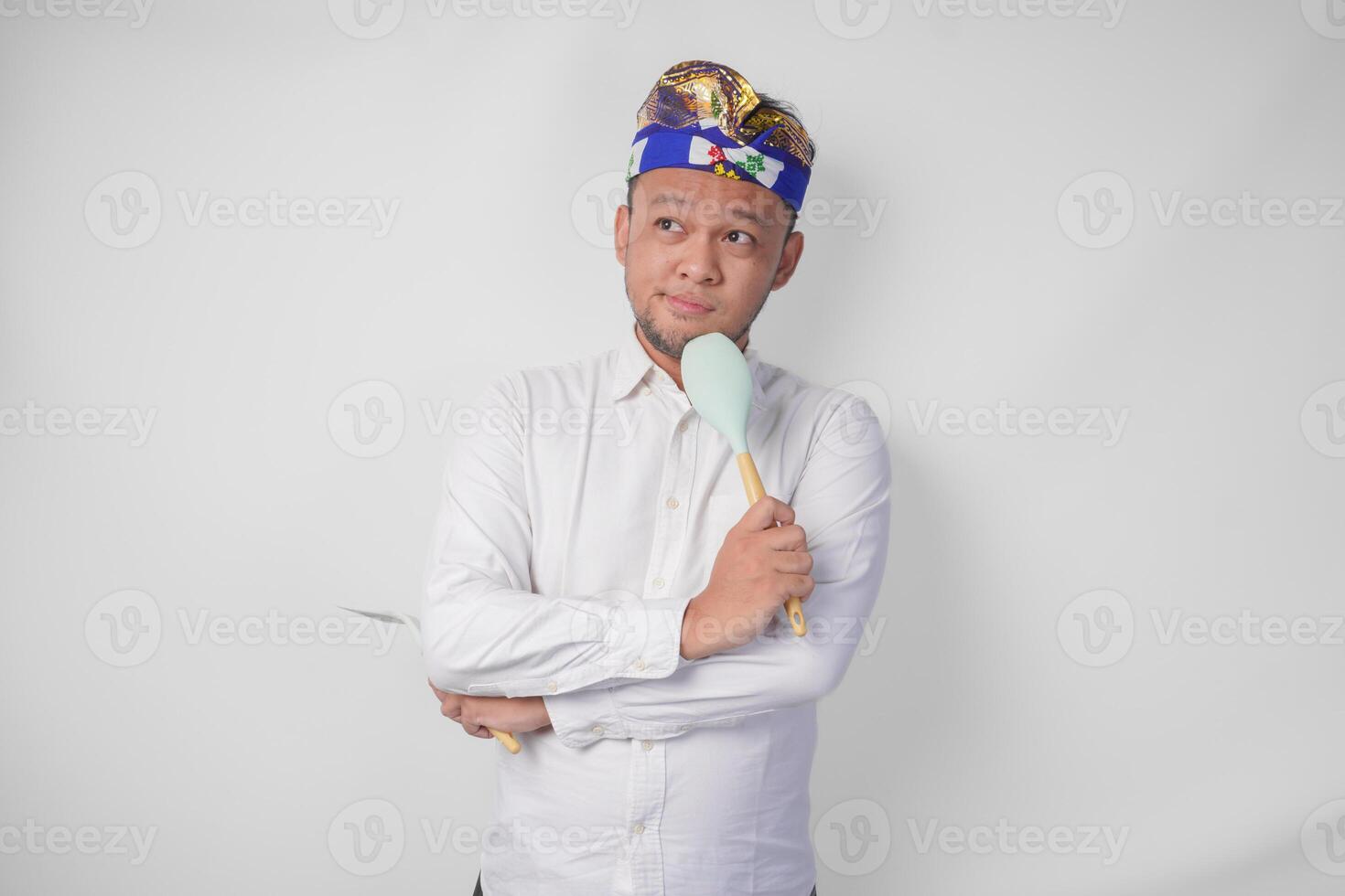 Thoughtful young Balinese man in white shirt and traditional headdress holding spatula and kitchen cooking utensils while thinking hard what food menu to cook photo