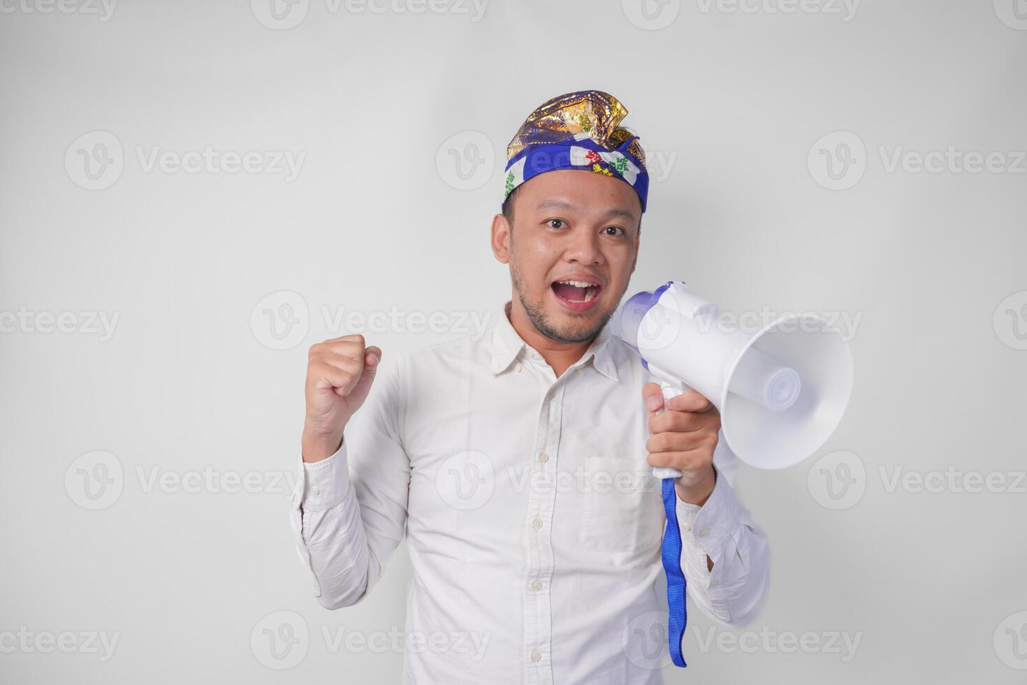 Energetic Balinese man in white shirt and traditional headdress shouting at megaphone while raising clenched fist, isolated by white background photo