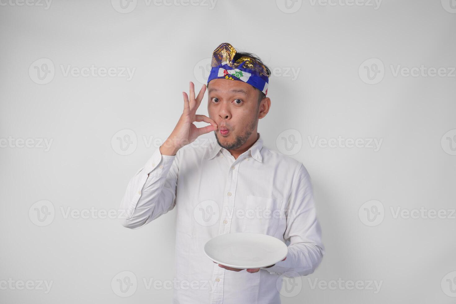 contento joven balinés hombre en blanco camisa y tradicional tocado participación un vacío plato con Copiar espacio mientras haciendo un delicioso mano gesto a Rápido cómo bueno el comida es foto