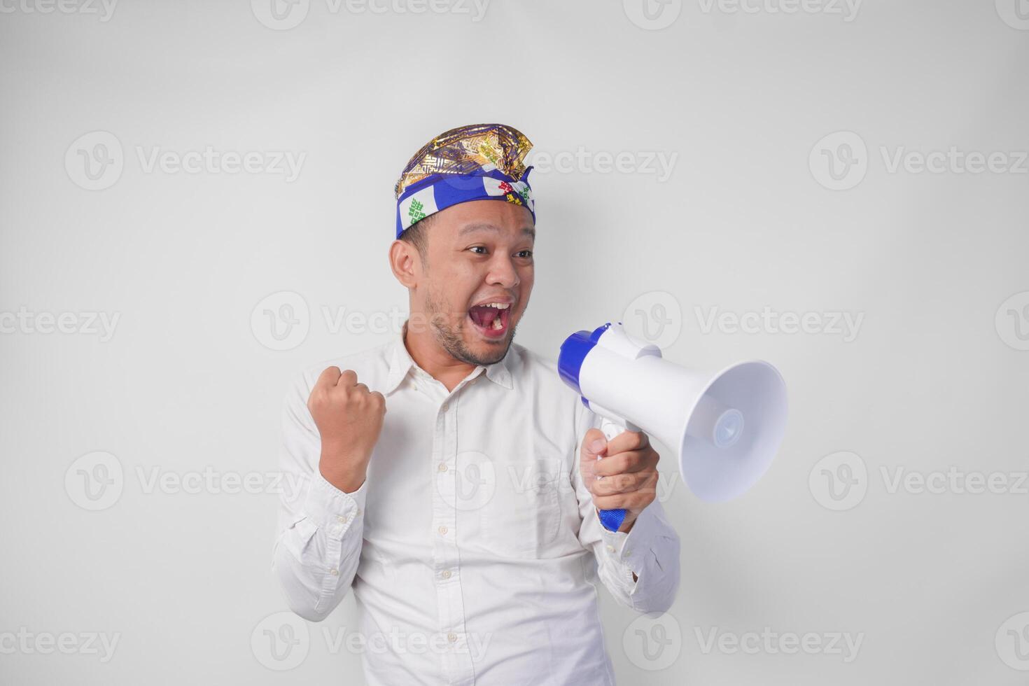 Overjoyed Balinese man in white shirt and traditional headdress shouting at megaphone feeling excited, isolated by white background photo