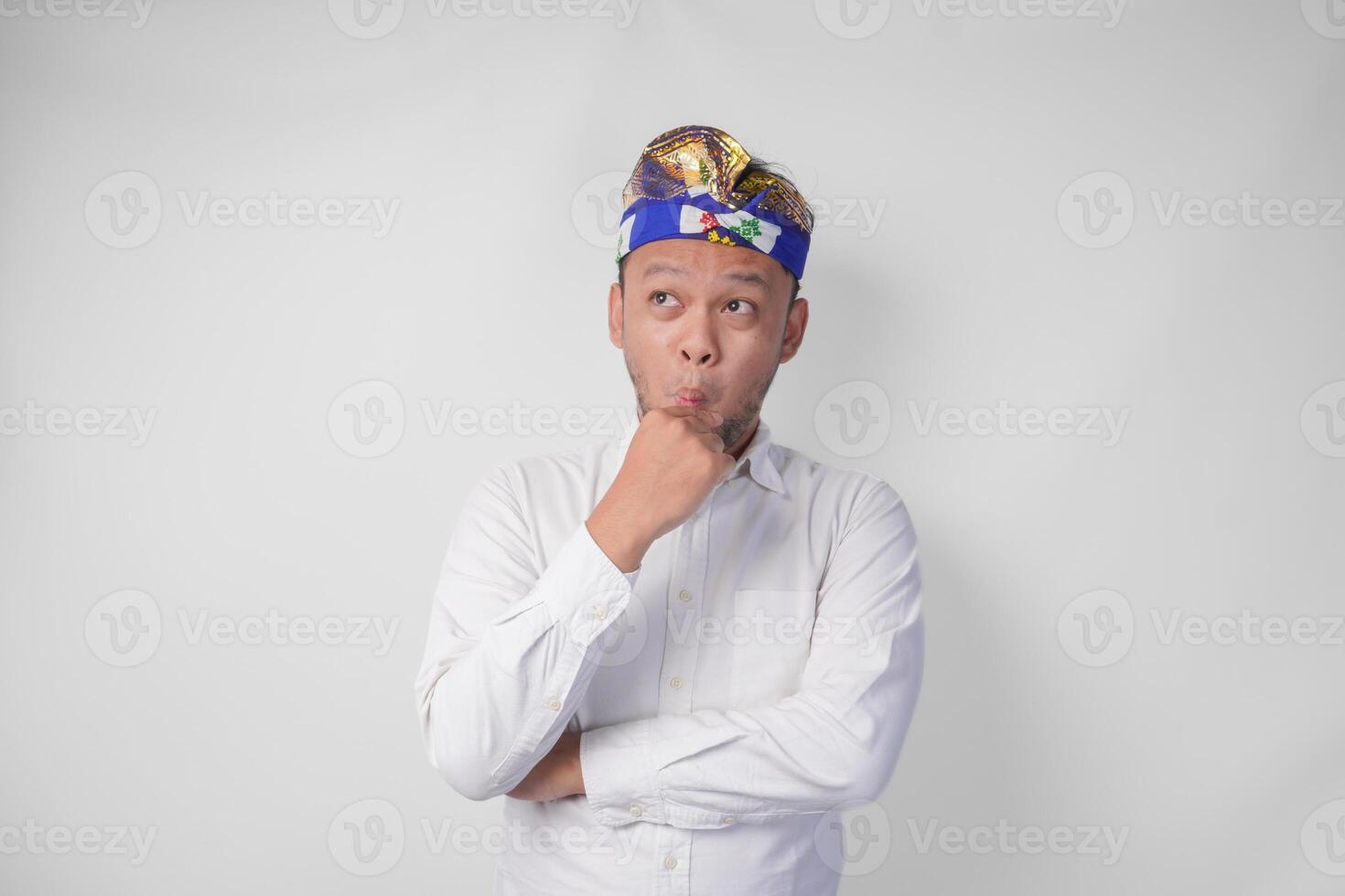 Confused Balinese man in traditional headdress called udeng thinking hard while touching chin with serious expression, isolated by white background photo