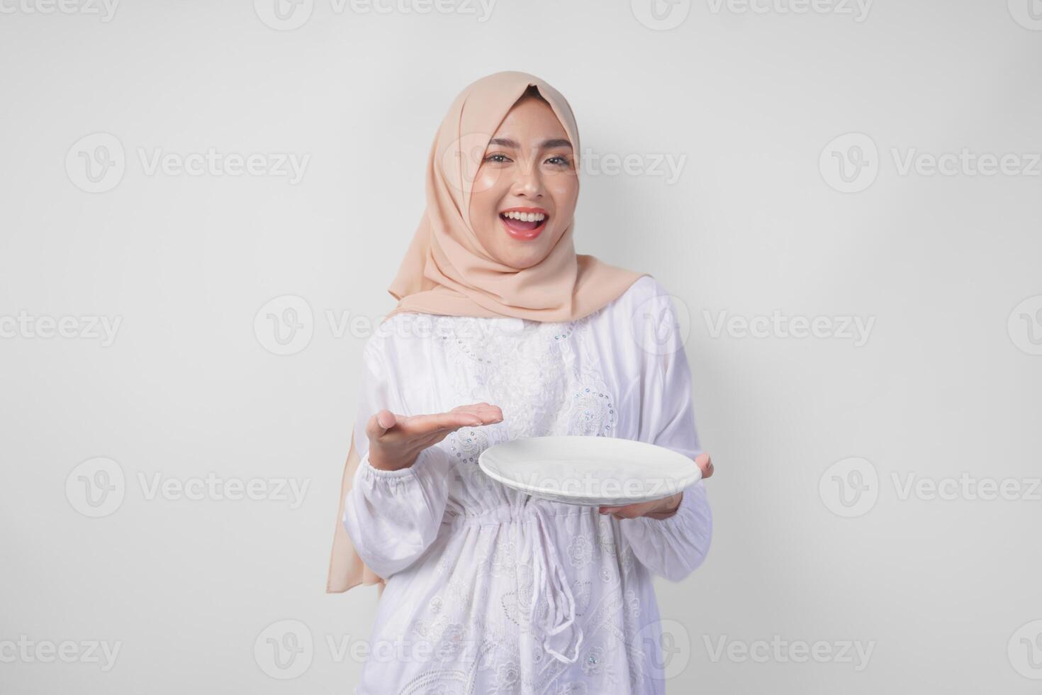 Smiling young Asian Muslim woman in a veil hijab pointing to an empty plate with copy space over it, presenting food menu for iftar. Ramadan concept photo