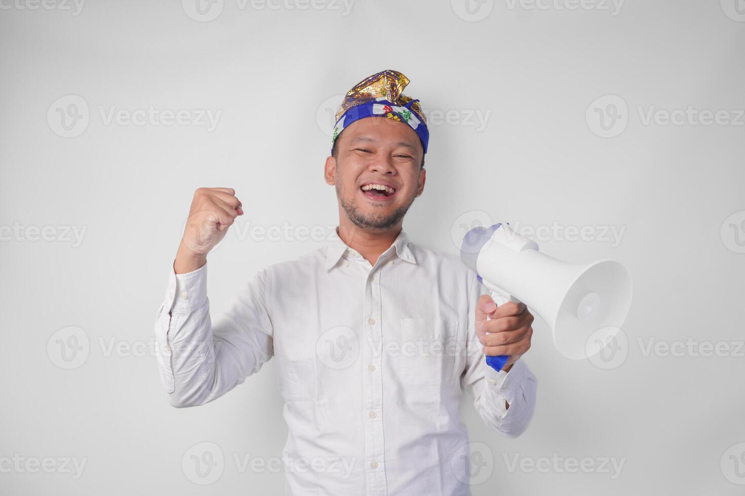 energético balinés hombre en blanco camisa y tradicional tocado gritos a megáfono mientras levantamiento apretado puño, aislado por blanco antecedentes foto