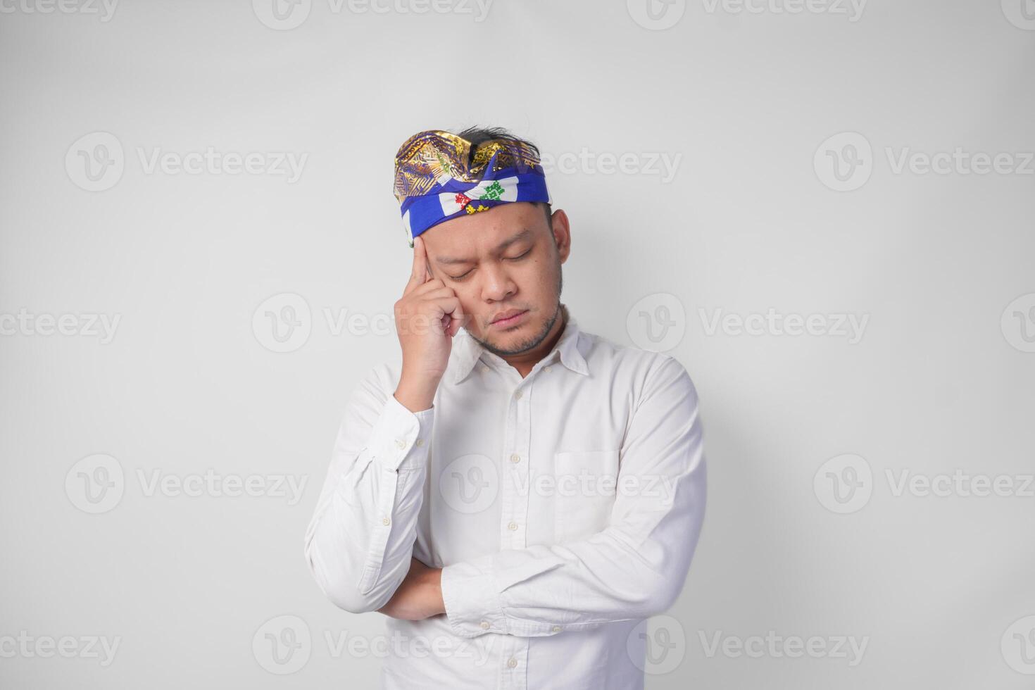Confused Balinese man in traditional headdress called udeng thinking hard while touching chin with serious expression, isolated by white background photo