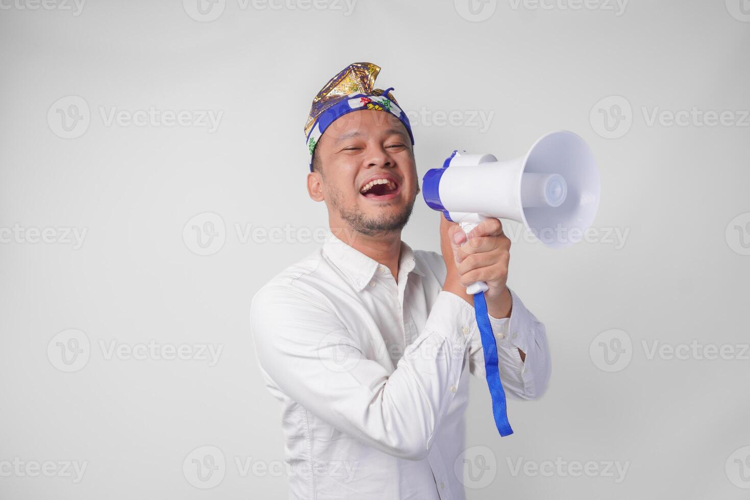 Energetic Balinese man in white shirt and traditional headdress shouting at megaphone while raising clenched fist, isolated by white background photo