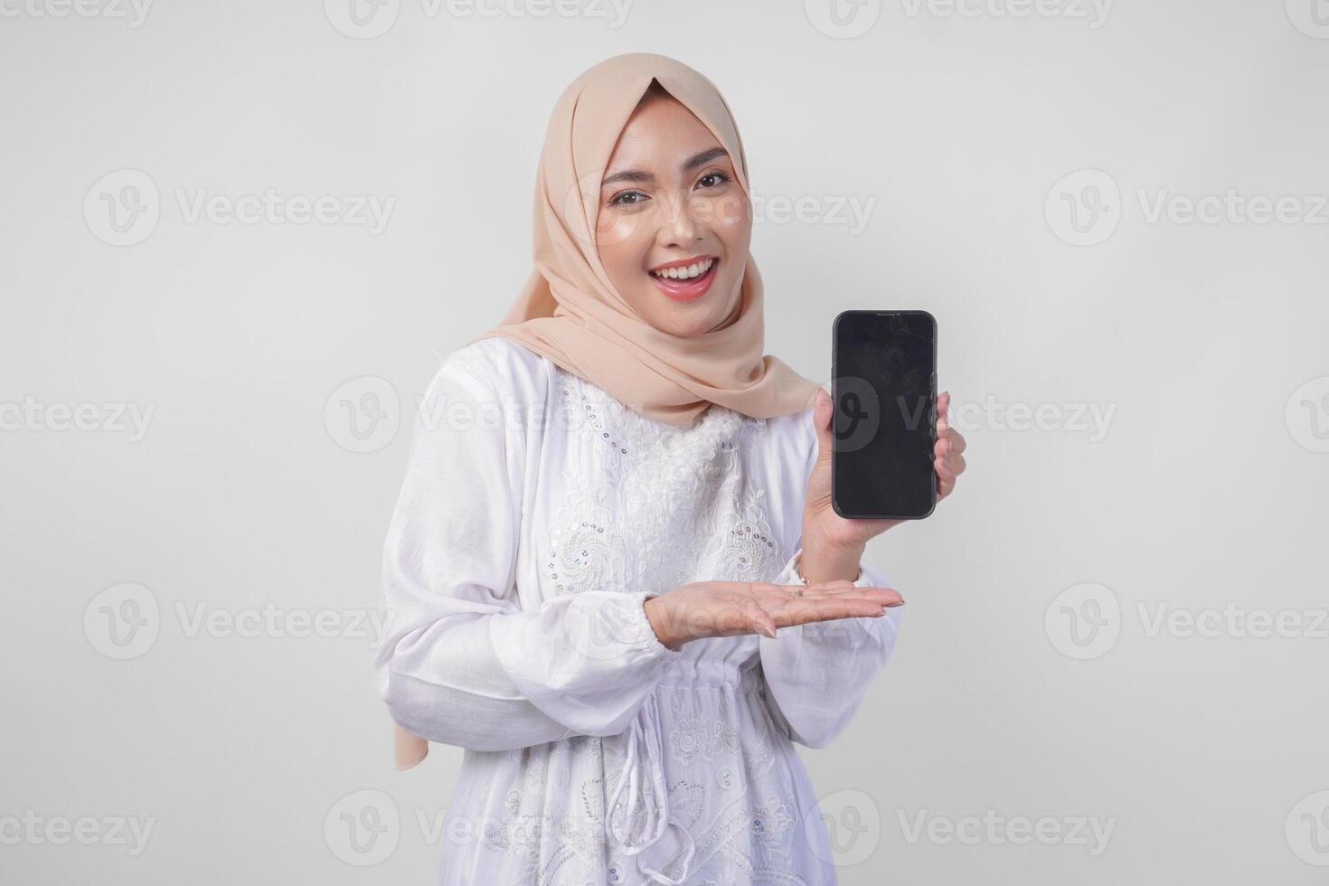 retrato de joven asiático musulmán mujer vistiendo blanco vestir y hijab sonriente felizmente, señalando a el blanco pantalla Copiar espacio en su teléfono y demostración pantalla a el cámara foto