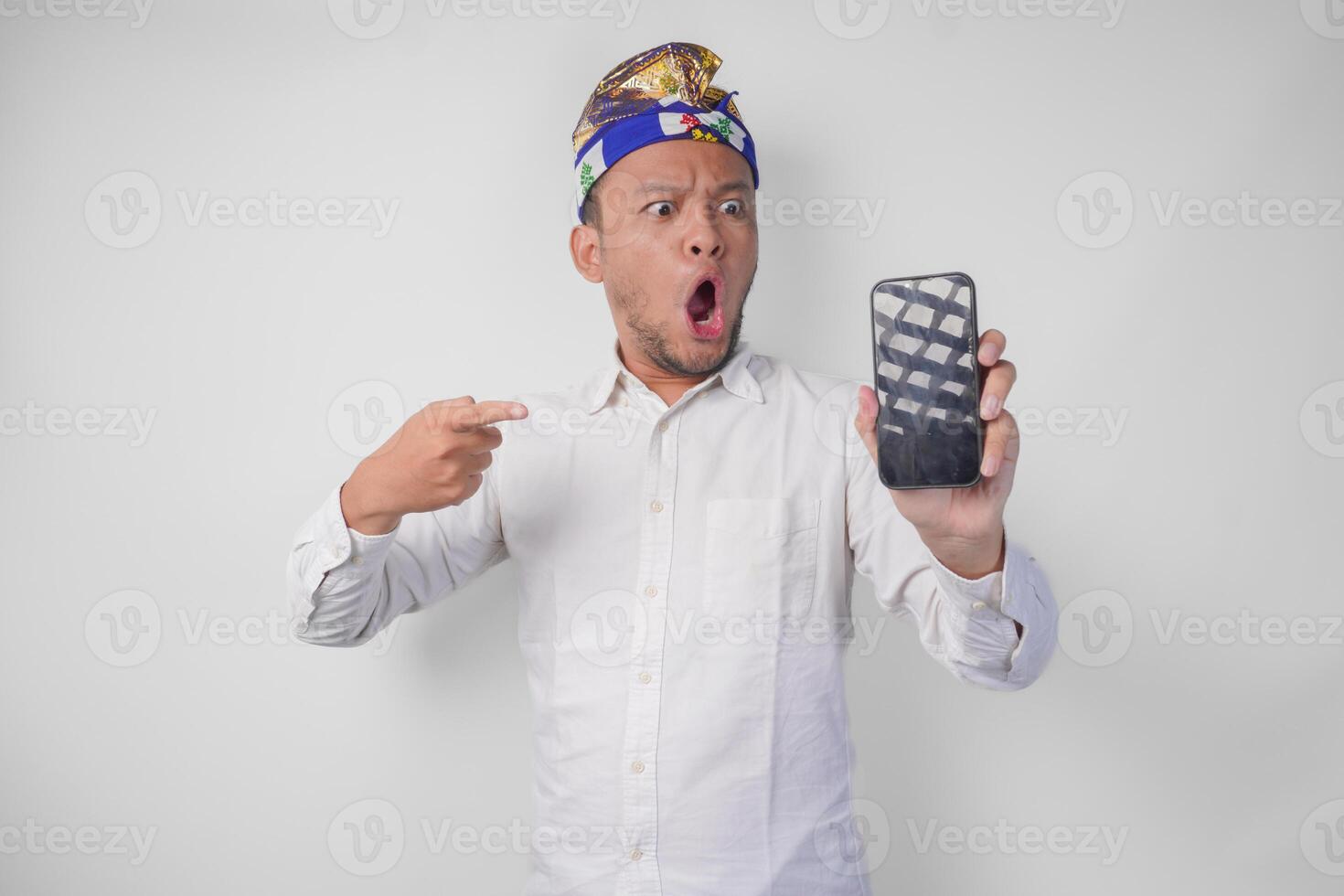 sorprendido joven balinés hombre vistiendo blanco camisa y tradicional tocado con conmocionado expresión en cara mientras señalando a su teléfono inteligente, presentación blanco pantalla Copiar espacio foto