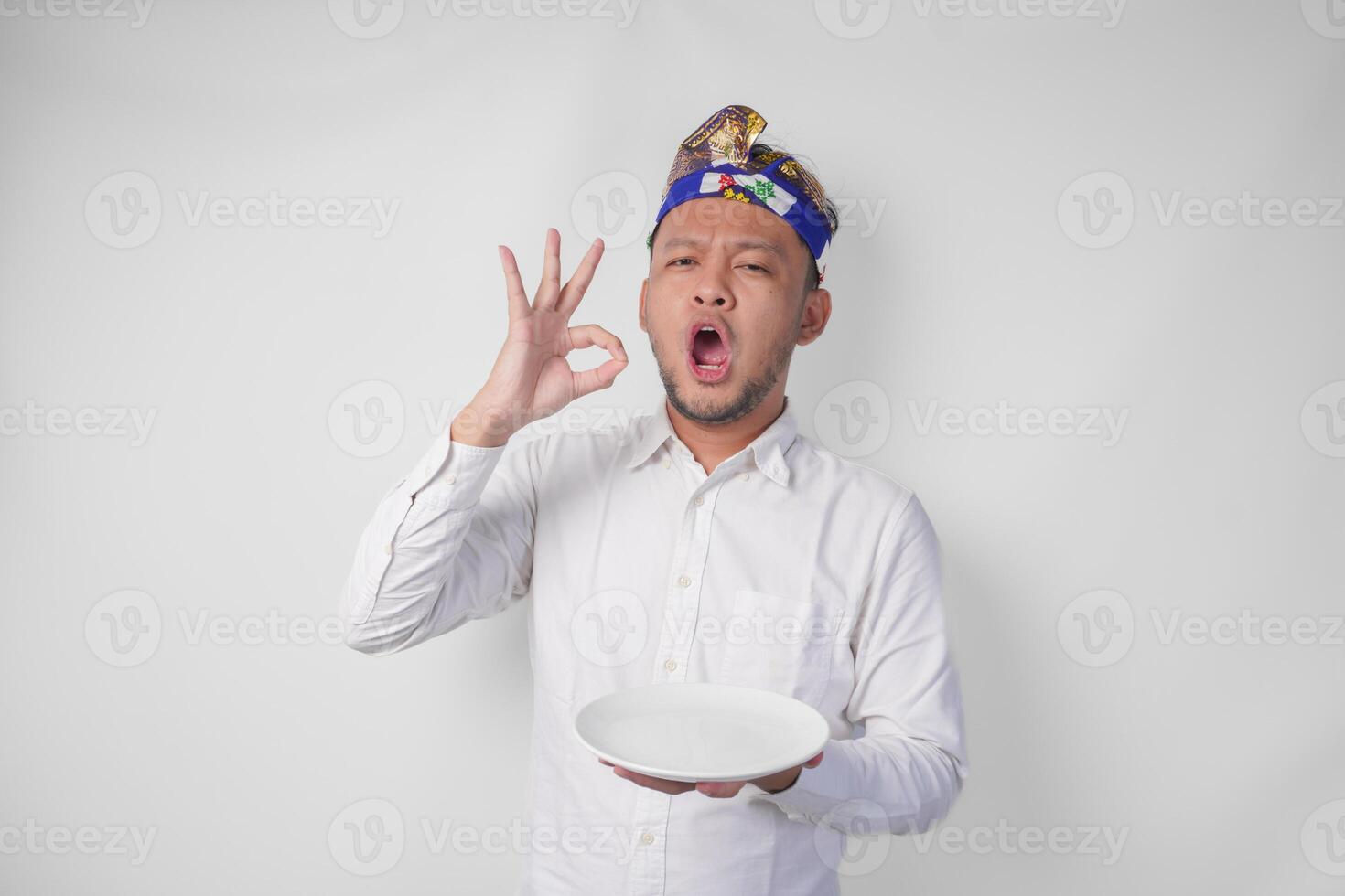 contento joven balinés hombre en blanco camisa y tradicional tocado participación un vacío plato con Copiar espacio mientras haciendo un delicioso mano gesto a Rápido cómo bueno el comida es foto