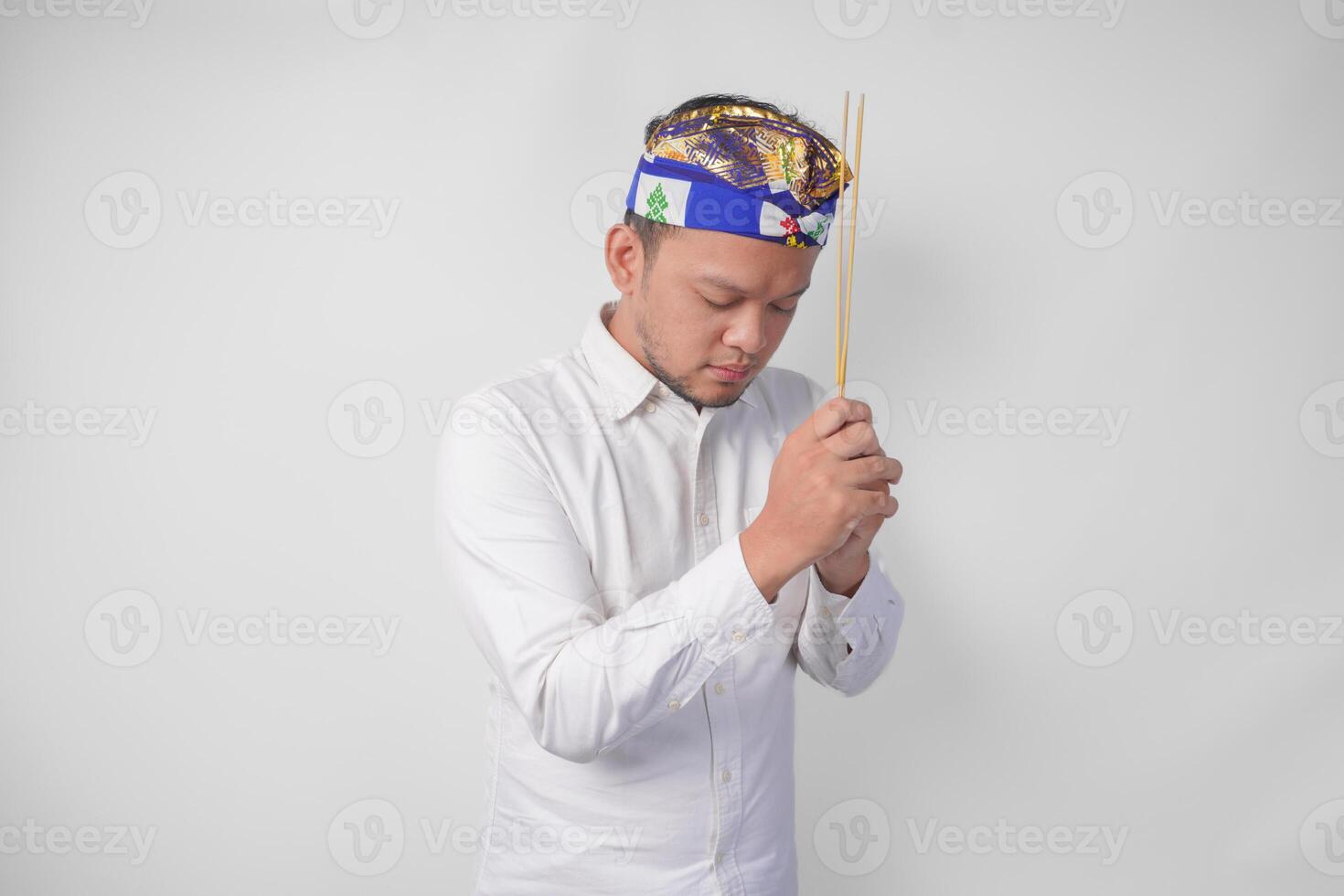 Balinese man wearing traditional headdress called udeng doing paying respect gesture while holding praying incense on isolated white background photo