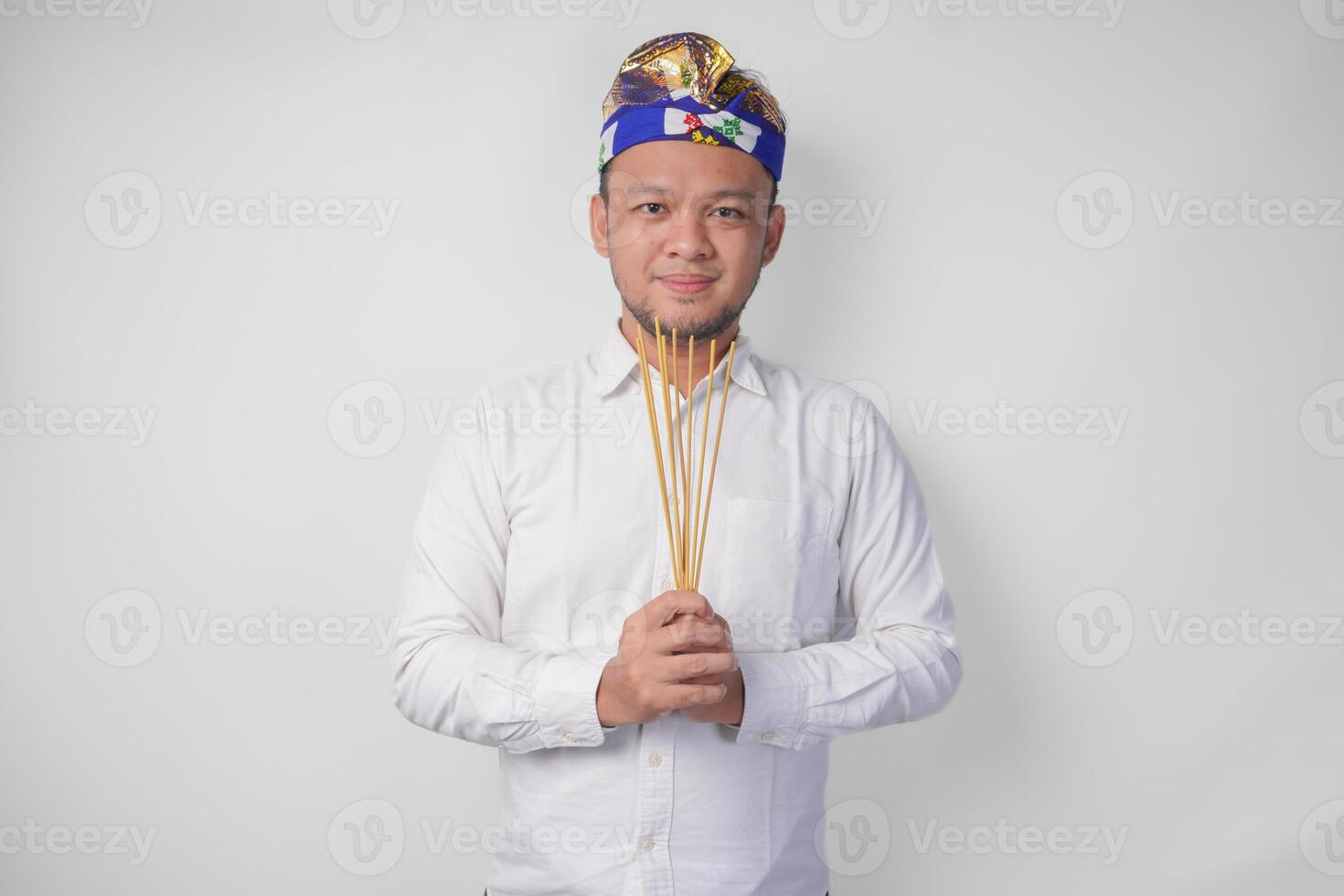 Balinese man wearing traditional headdress called udeng doing paying respect gesture while holding praying incense on isolated white background photo