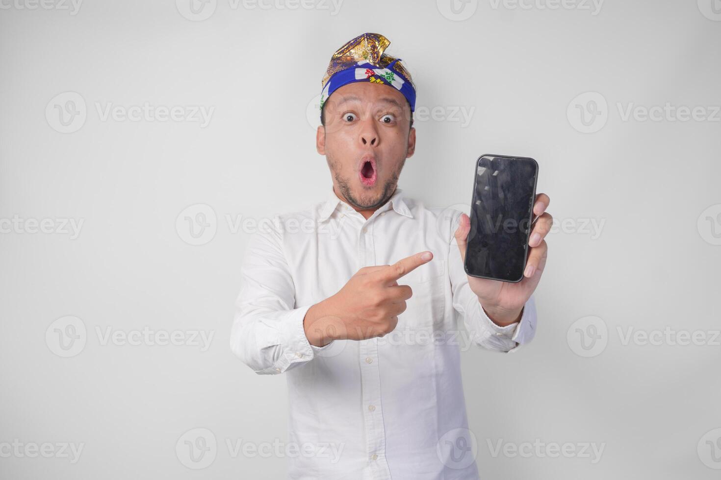 Surprised young Balinese man wearing white shirt and traditional headdress with shocked expression on face while pointing to his smartphone, presenting blank screen copy space photo