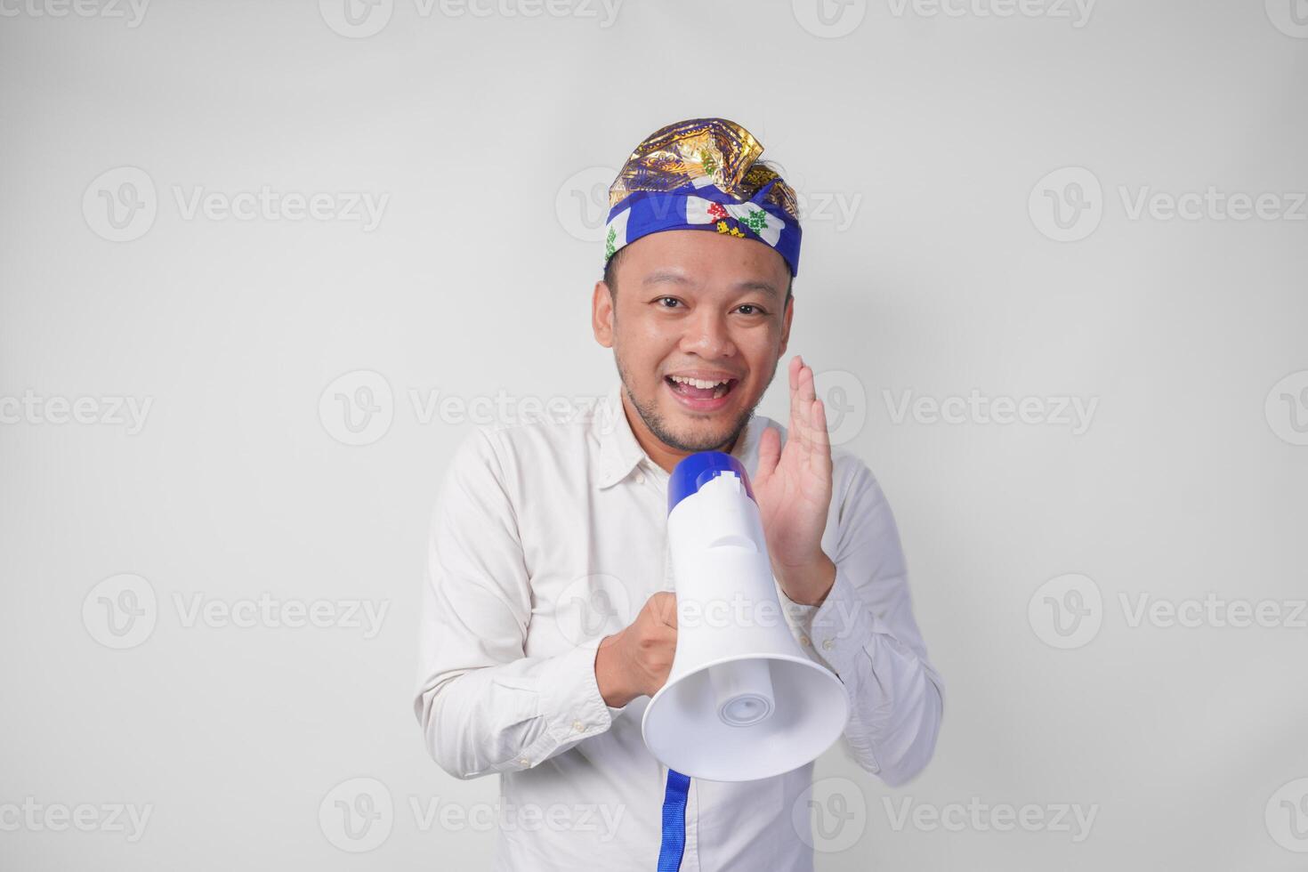 Energetic Balinese man in white shirt and traditional headdress shouting at megaphone while raising clenched fist, isolated by white background photo