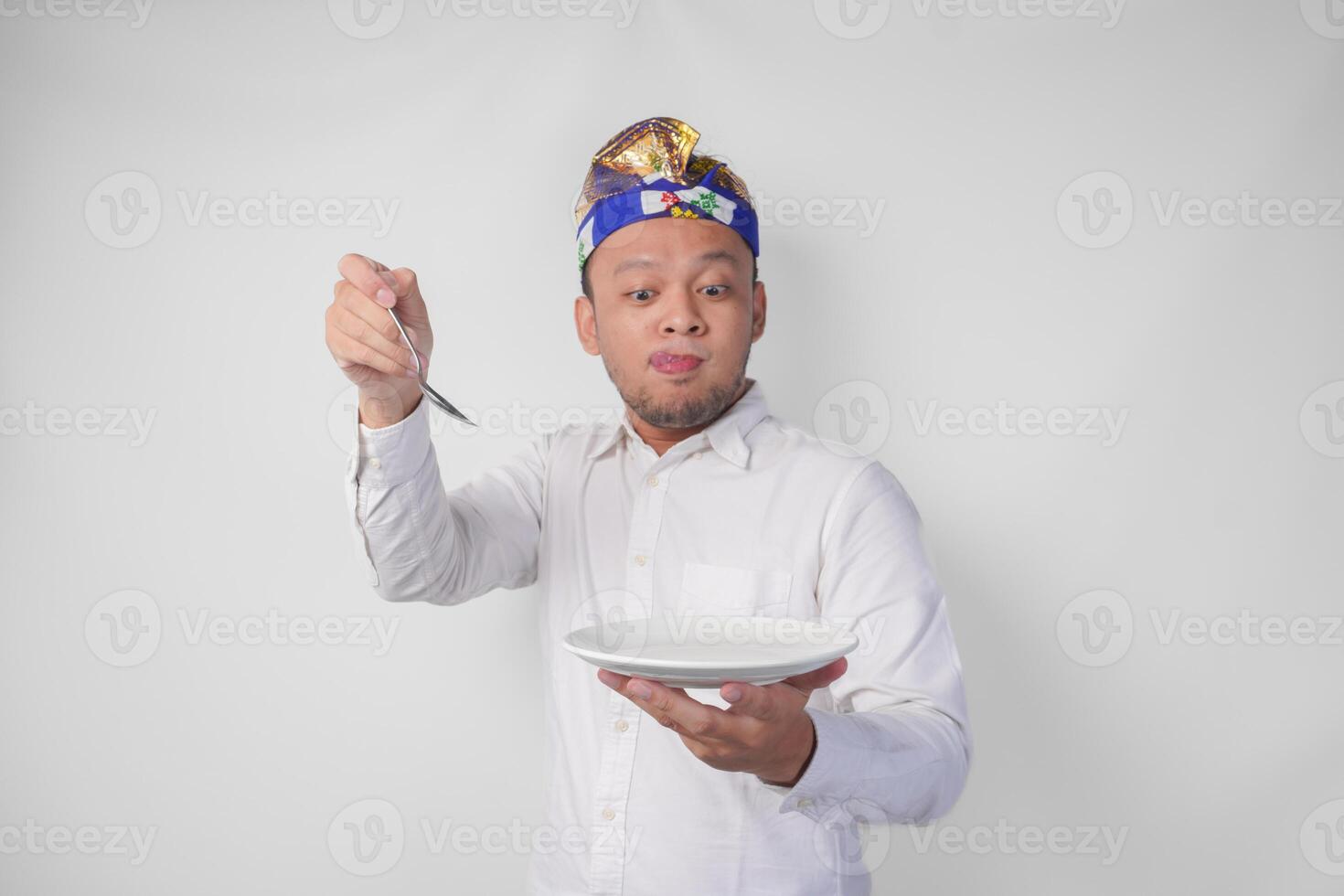 hambriento joven balinés hombre en blanco camisa y tradicional tocado participación un vacío plato con Copiar espacio y cuchara mientras haciendo un gracioso expresión foto