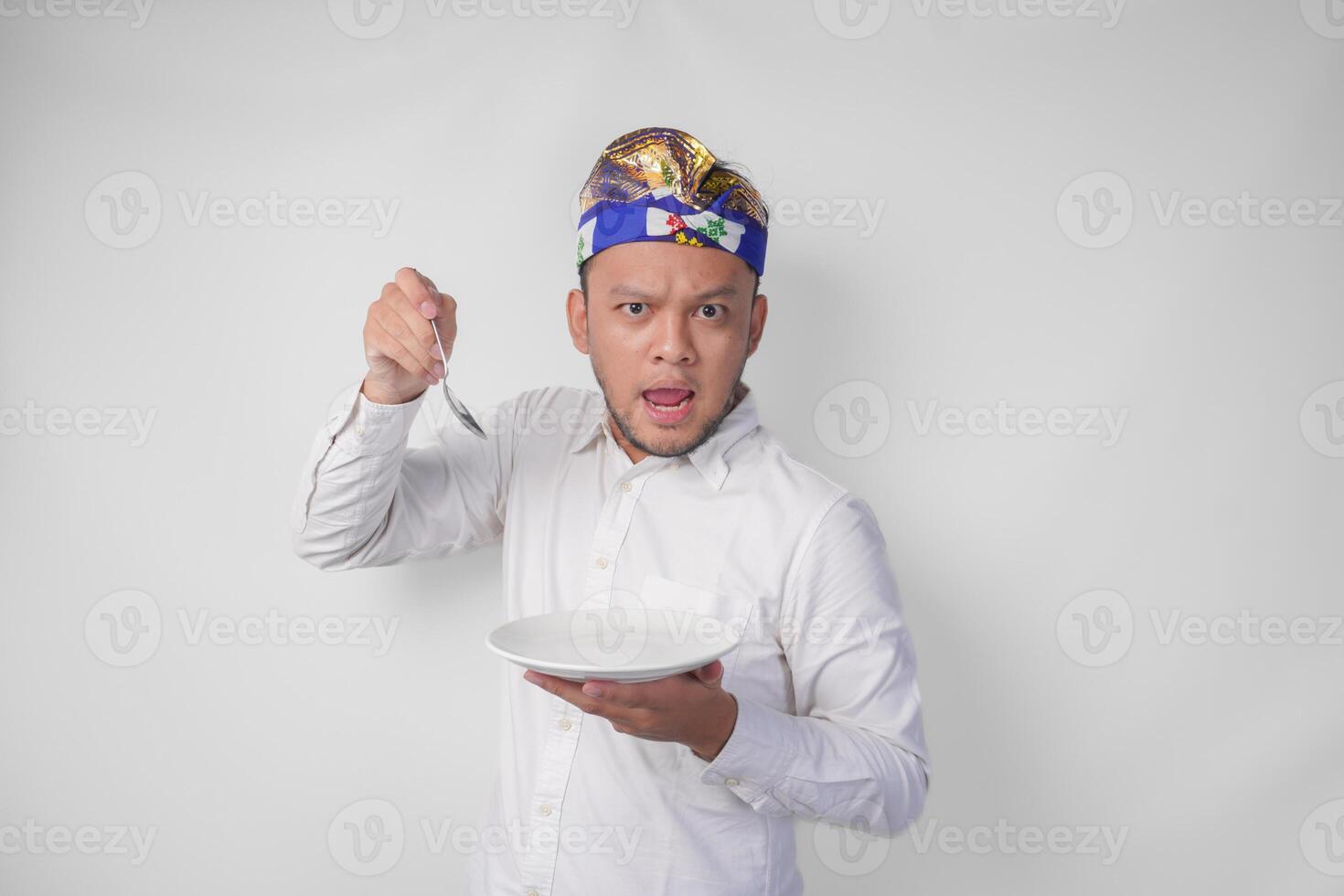 Hungry young Balinese man in white shirt and traditional headdress holding an empty plate with copy space and spoon while making a funny expression photo