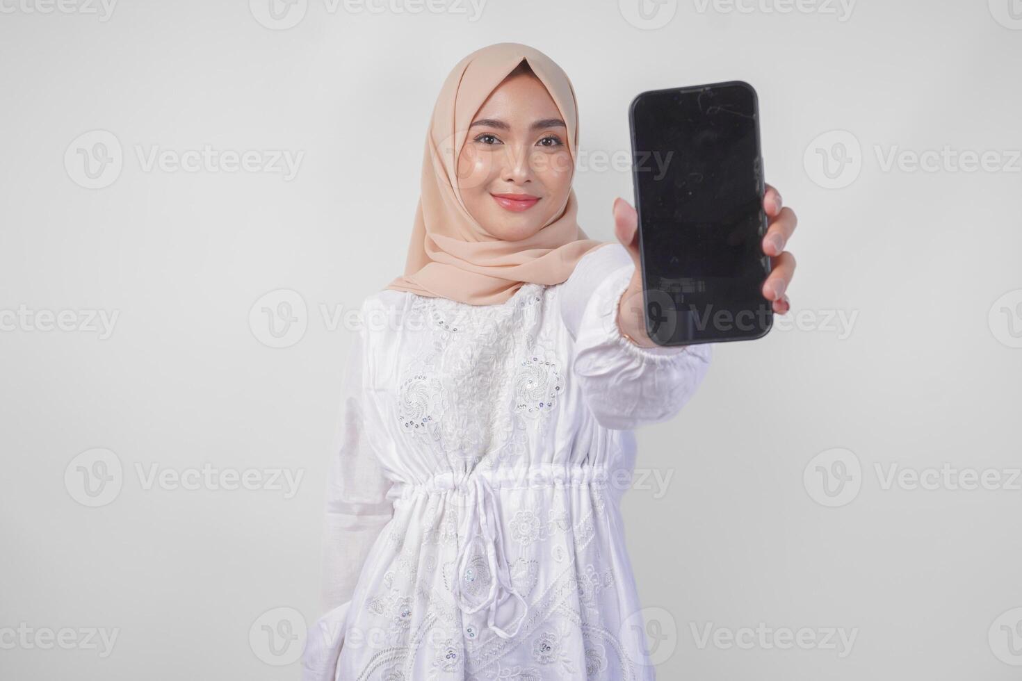 Portrait of young Asian Muslim woman wearing white dress and hijab smiling happily, pointing to the blank screen copy space on her phone and showing screen to the camera photo