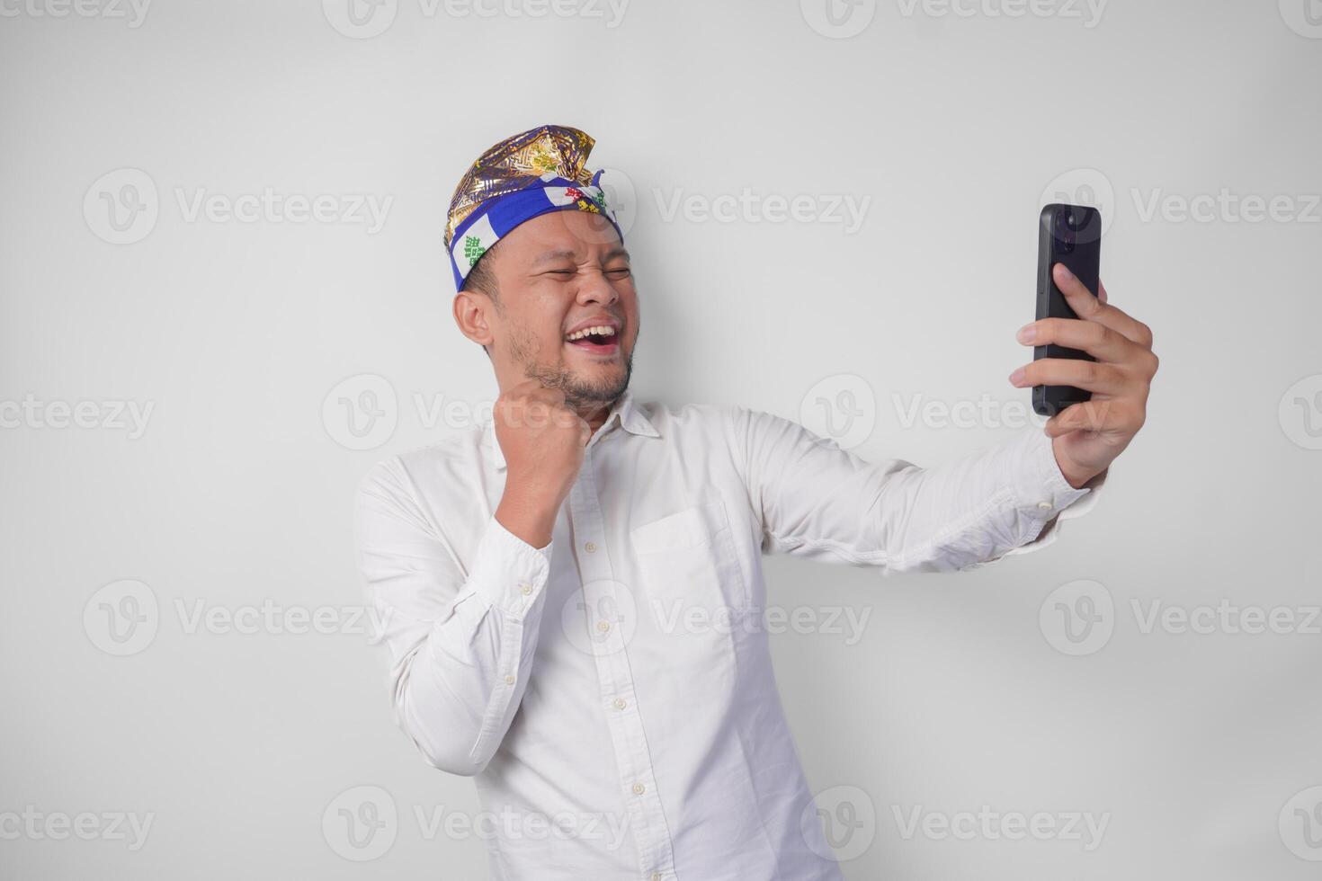 Young Balinese man in white shirt and traditional headdress doing call with family or friend feeling overjoyed and excited photo