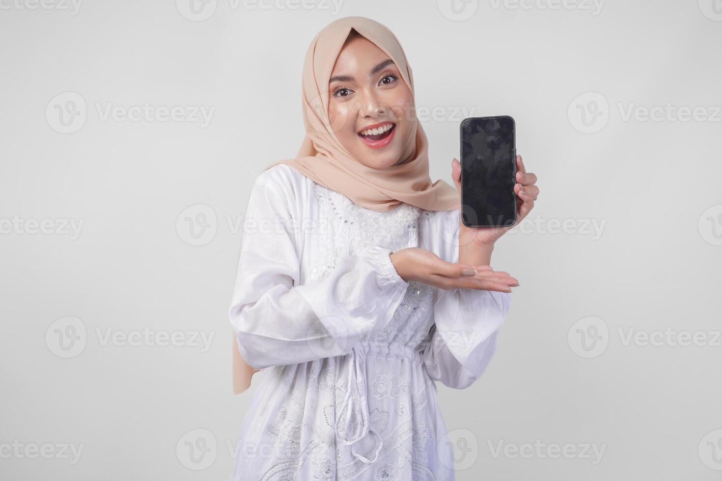 Portrait of young Asian Muslim woman wearing white dress and hijab smiling happily, pointing to the blank screen copy space on her phone and showing screen to the camera photo