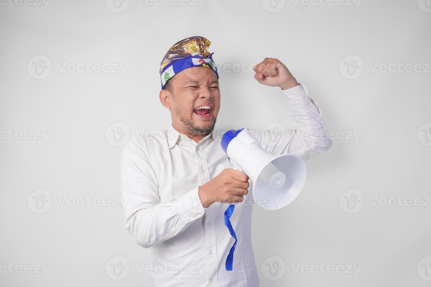 Energetic Balinese man in white shirt and traditional headdress shouting at megaphone while raising clenched fist, isolated by white background photo