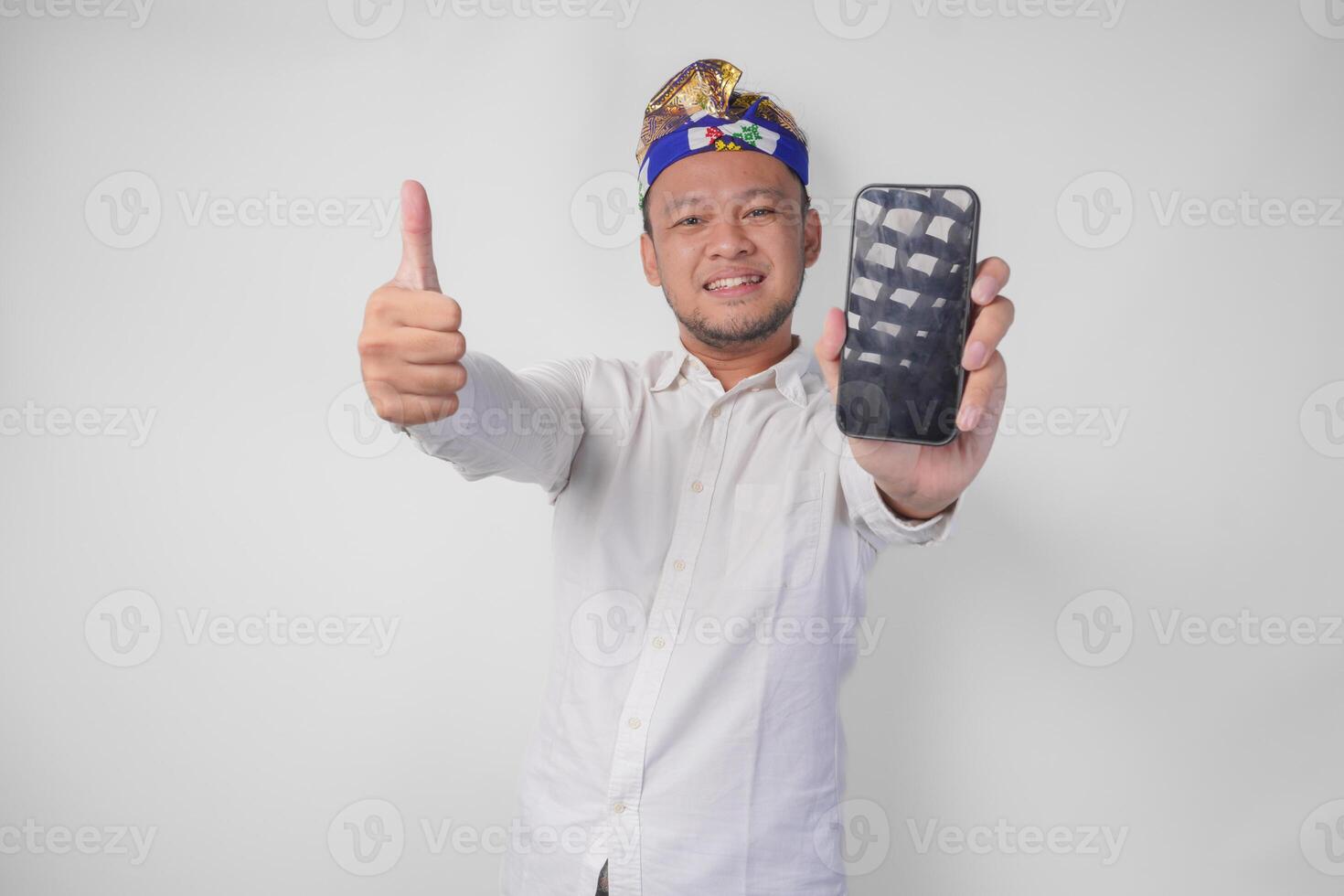 Young Balinese man wearing white shirt and traditional headdress presenting blank screen copy space on his phone while raising thumb up as an okay, good, nice gesture photo