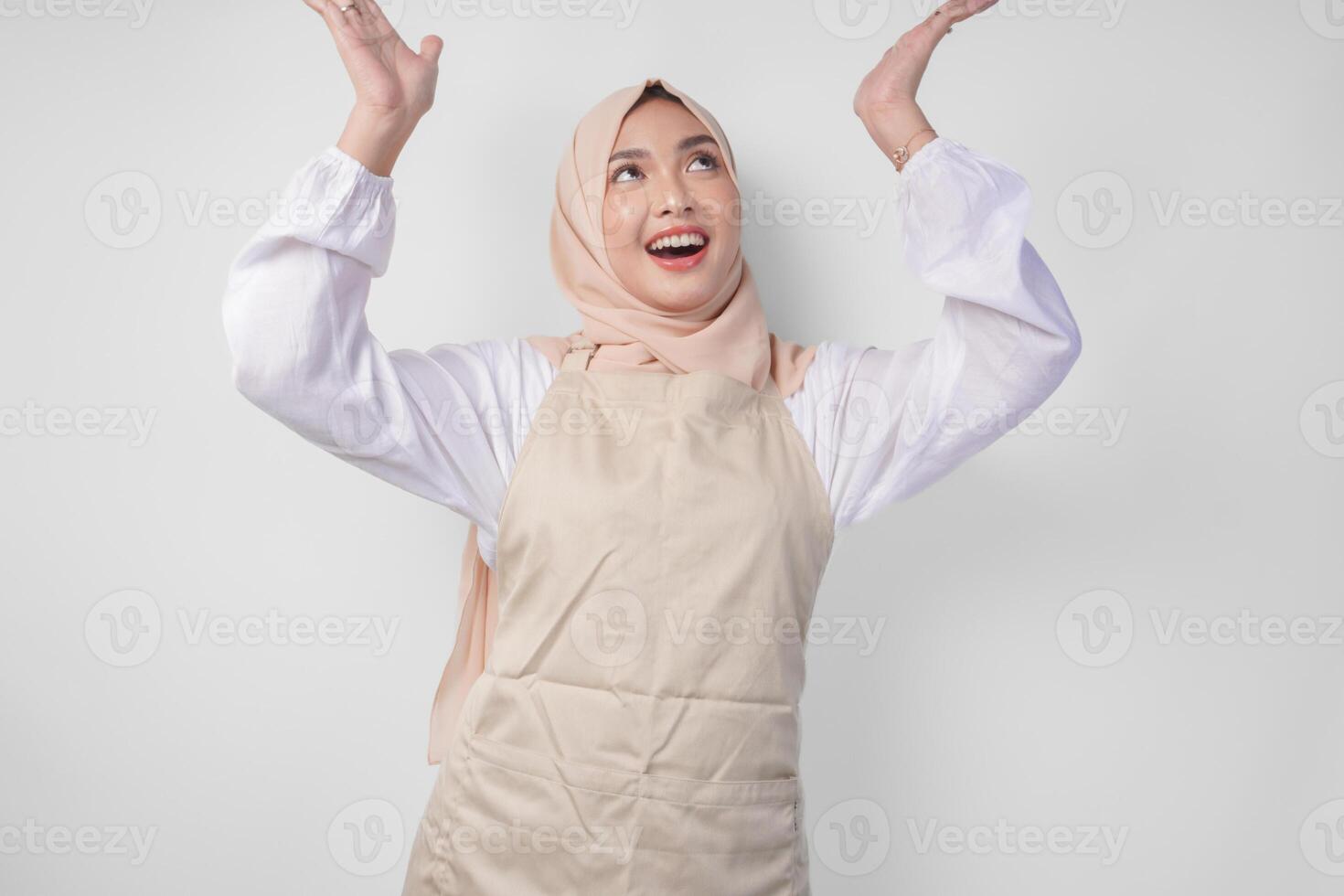 Overjoyed Asian woman wearing hijab and cream apron pointing up to the copy space above, standing over isolated white background photo