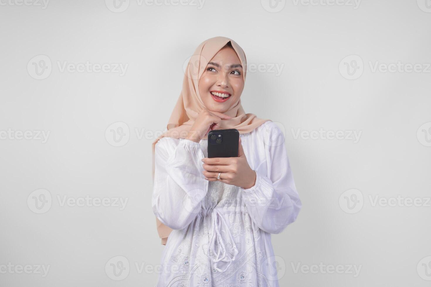 Thoughtful young Asian Muslim woman wearing white dress and hijab holding phone while thinking and finding idea with happy expression, isolated on white background. Ramadan and Eid Mubarak concept photo