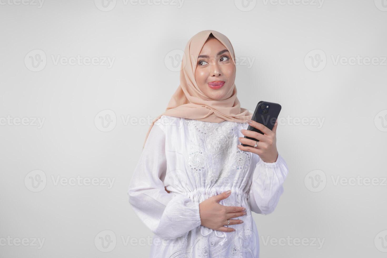 retrato de joven asiático musulmán mujer en blanco vestir y hijab sensación hambriento mientras mirando a su teléfono a orden sabroso comida después ayuno, aislado en blanco antecedentes foto