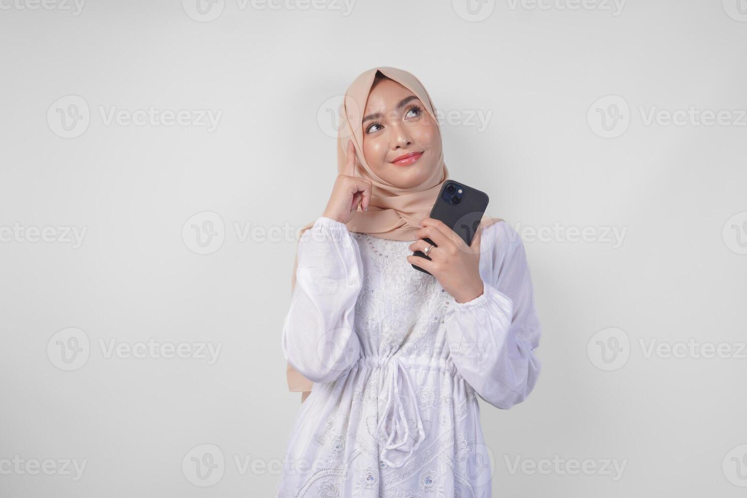 Thoughtful young Asian Muslim woman wearing white dress and hijab, using smartphone while holding her chin and thinking with serious expression over isolated white background photo