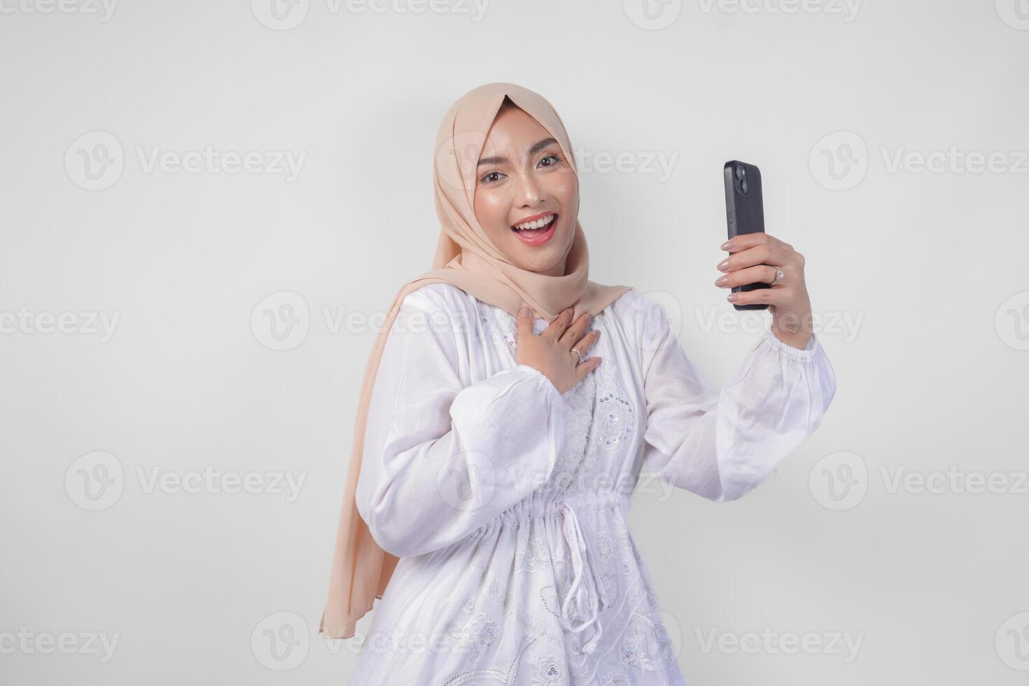 Portrait of Asian Muslim woman wearing white dress and hijab holding her smartphone to do a call to family or friend, isolated by white background. Ramadhan and Eid Mubarak concept photo