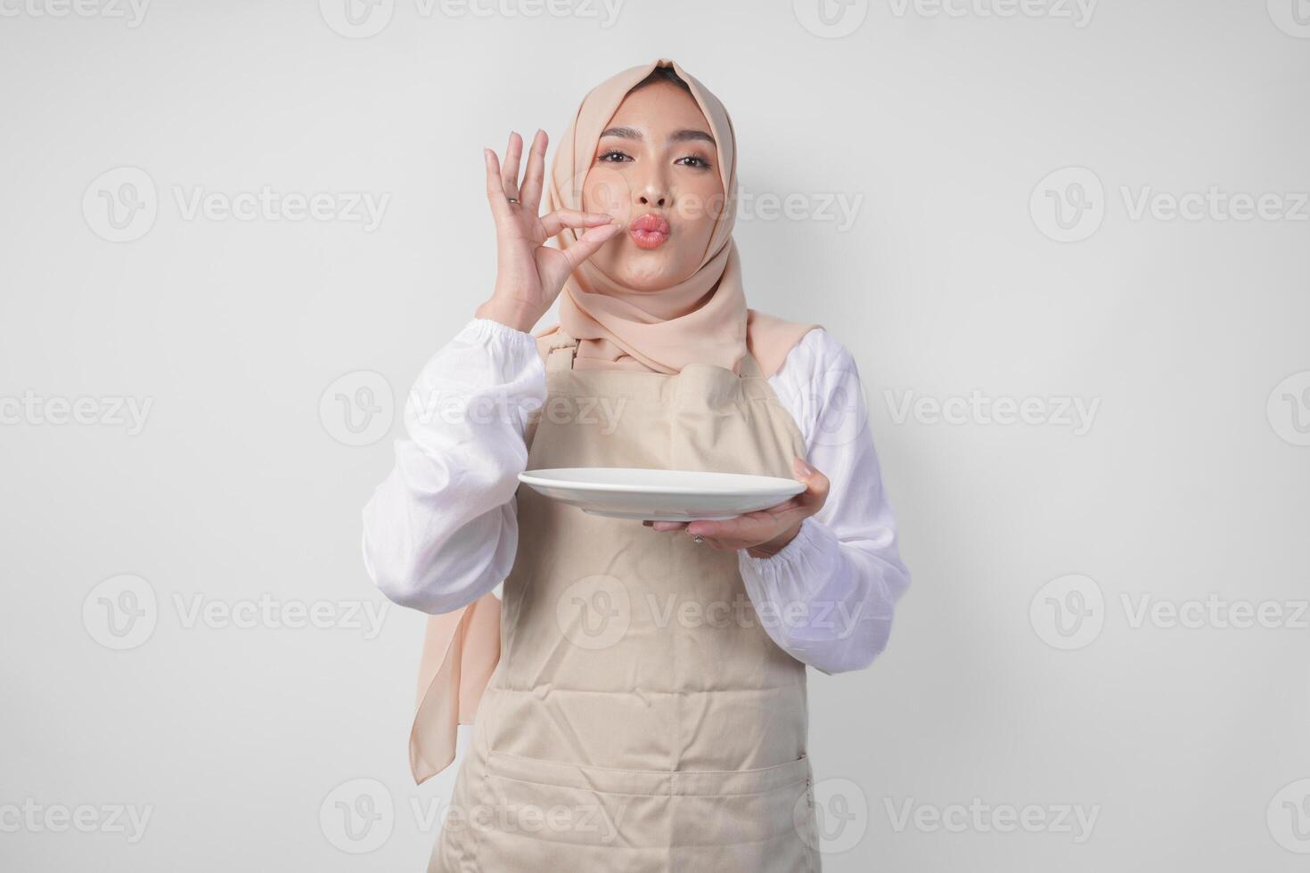 Happy young Asian Muslim woman in hijab and cream apron holding an empty plate with copy space while making a delicious hand gesture to express how good the food is photo