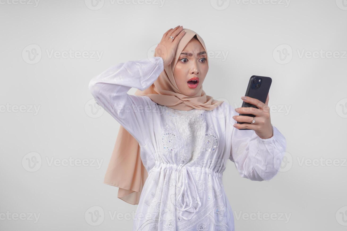 Young Asian Muslim woman wearing white dress and hijab showing shocked expression on her face while holding smartphone, surprised after reading news or gossip, isolated by white background photo