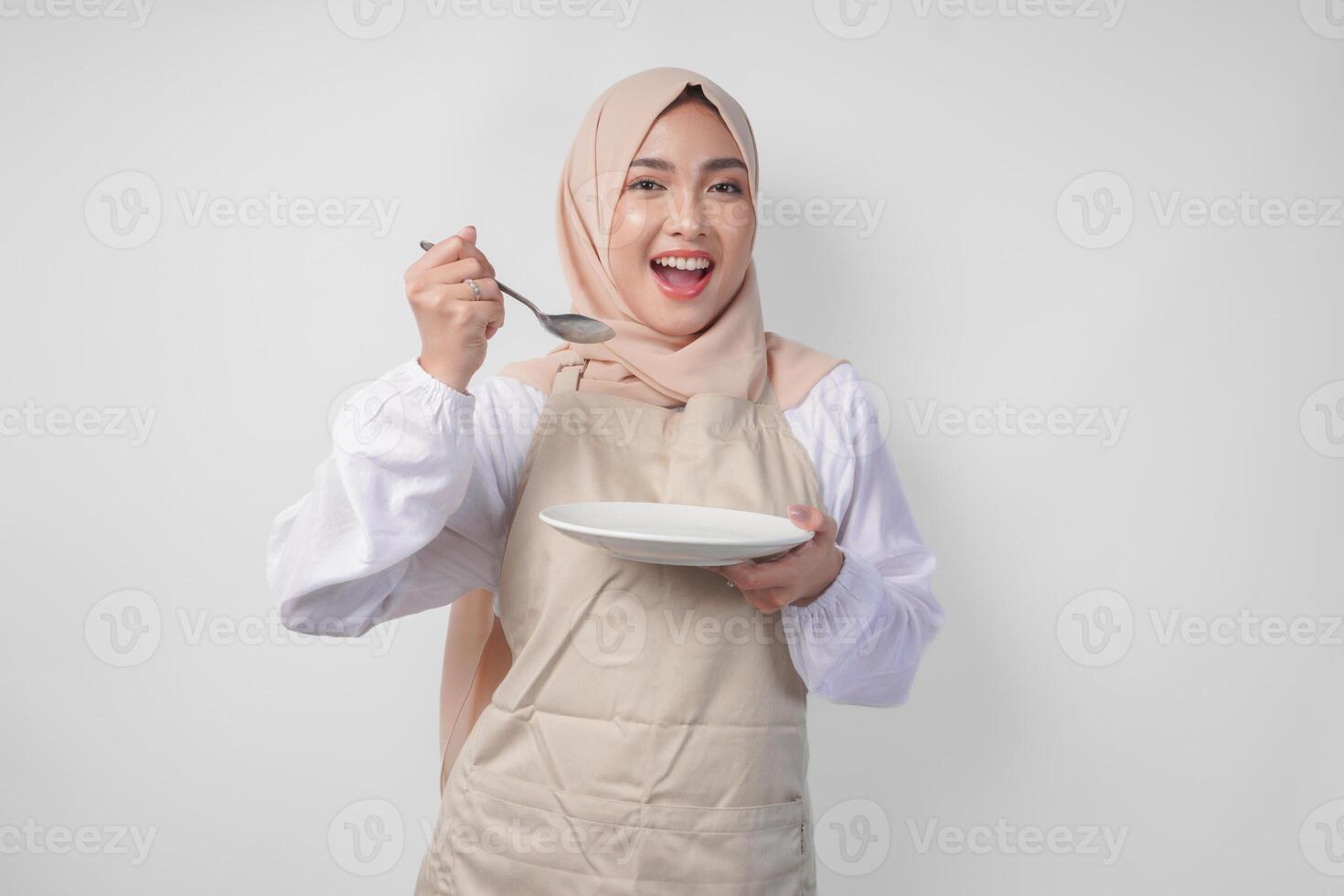 Hungry young Asian Muslim woman in hijab and cream apron holding spoon and eating from an empty plate with copy space over it. Ramadan concept photo