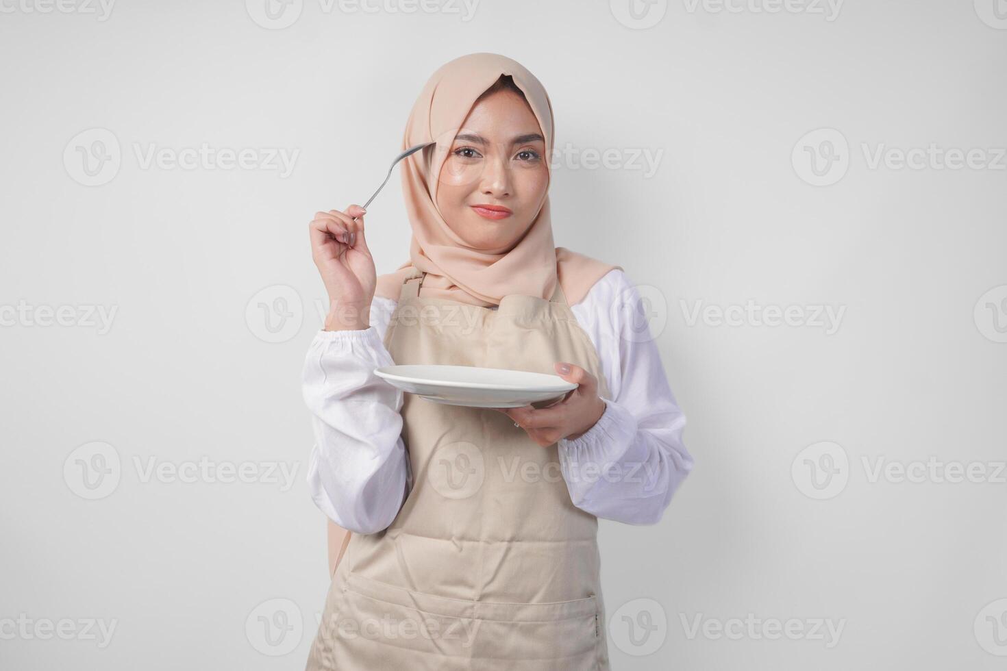 Confused young Asian Muslim woman in hijab and cream apron holding spoon and empty plate with copy space over it, thinking hard what food menu to eat for iftar. Ramadan concept photo