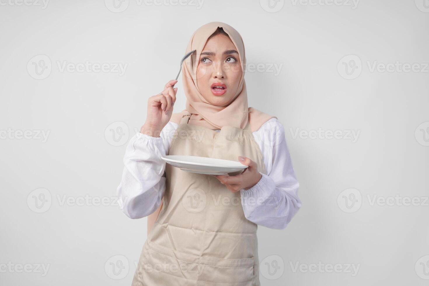 Confused young Asian Muslim woman in hijab and cream apron holding spoon and empty plate with copy space over it, thinking hard what food menu to eat for iftar. Ramadan concept photo