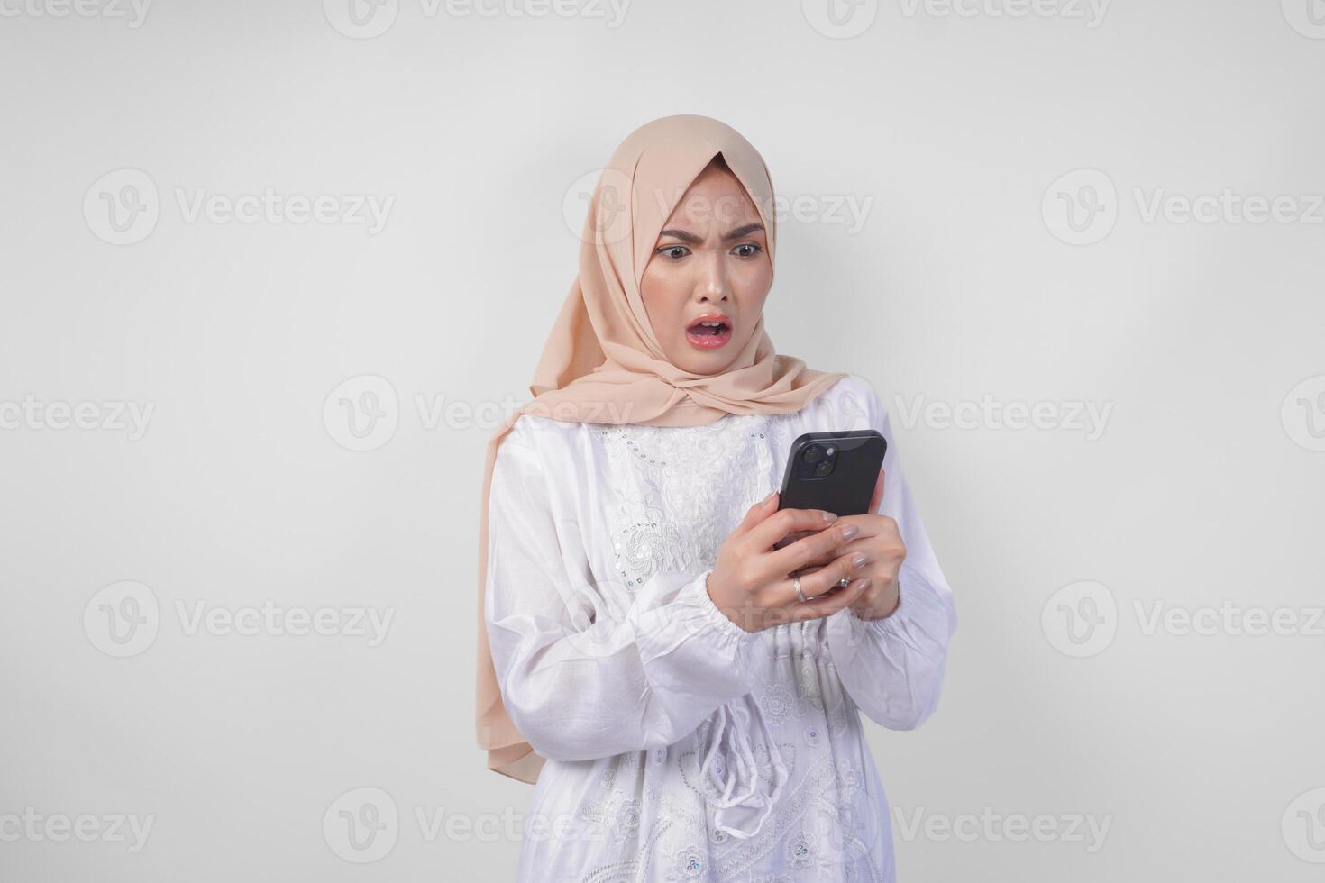 joven asiático musulmán mujer vistiendo blanco vestir y hijab demostración conmocionado expresión en su cara mientras participación teléfono inteligente, sorprendido después leyendo Noticias o chisme, aislado por blanco antecedentes foto