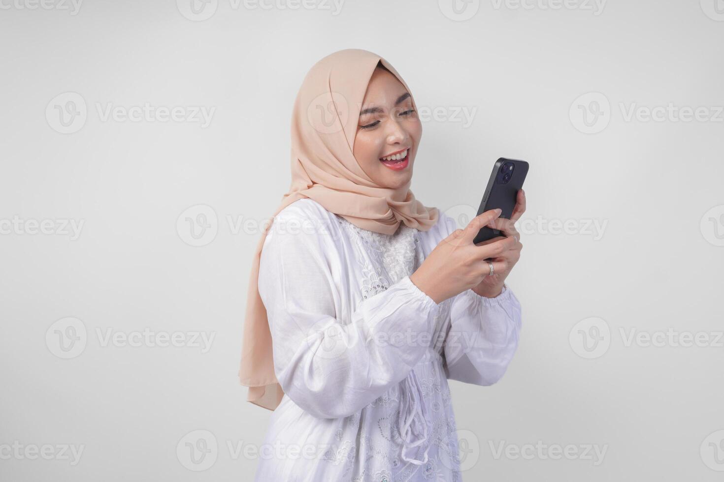 Excited Asian Muslim woman wearing white dress and hijab holding her smartphone while smiling cheerfully, isolated by white background. Ramadhan and Eid Mubarak concept photo