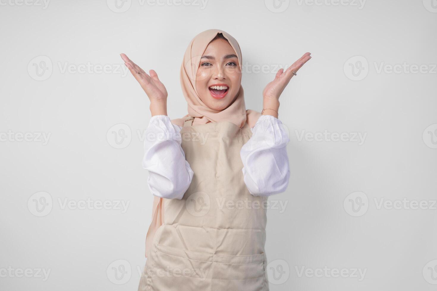 Overjoyed Asian woman wearing hijab and cream apron pointing up to the copy space above, standing over isolated white background photo