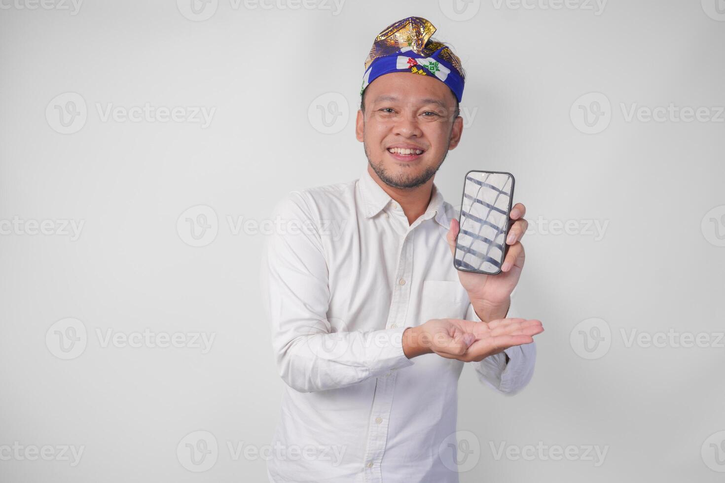 Smiling young Balinese man wearing white shirt and traditional headdress pointing to his smartphone, presenting blank screen copy space photo