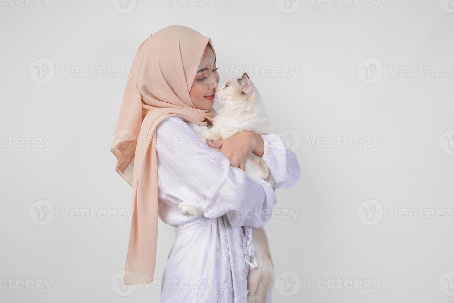 Portrait of a beautiful young Muslim woman wearing white dress and hijab, smiling and holding a white ragdoll cat over isolated white background photo