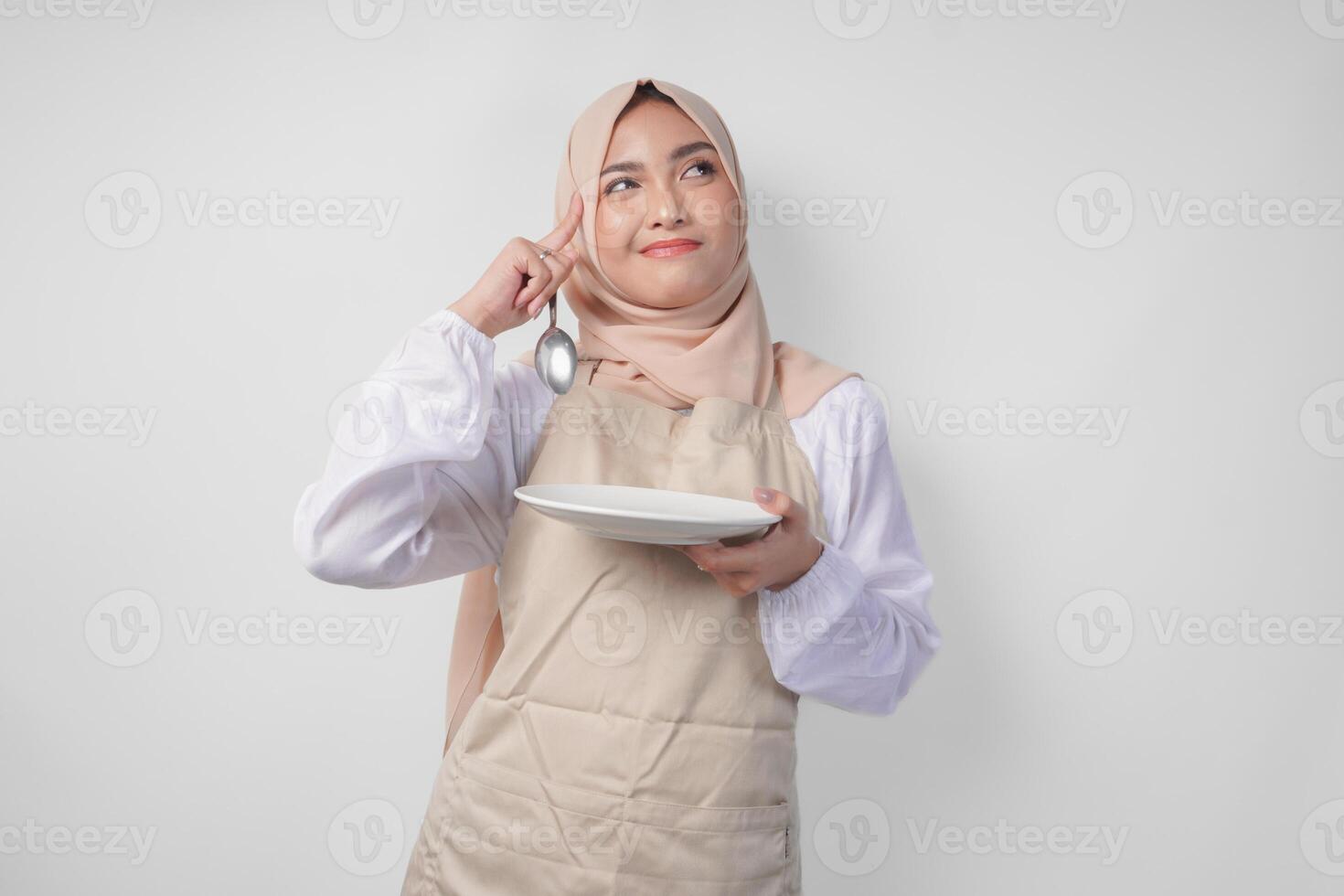 Confused young Asian Muslim woman in hijab and cream apron holding spoon and empty plate with copy space over it, thinking hard what food menu to eat for iftar. Ramadan concept photo